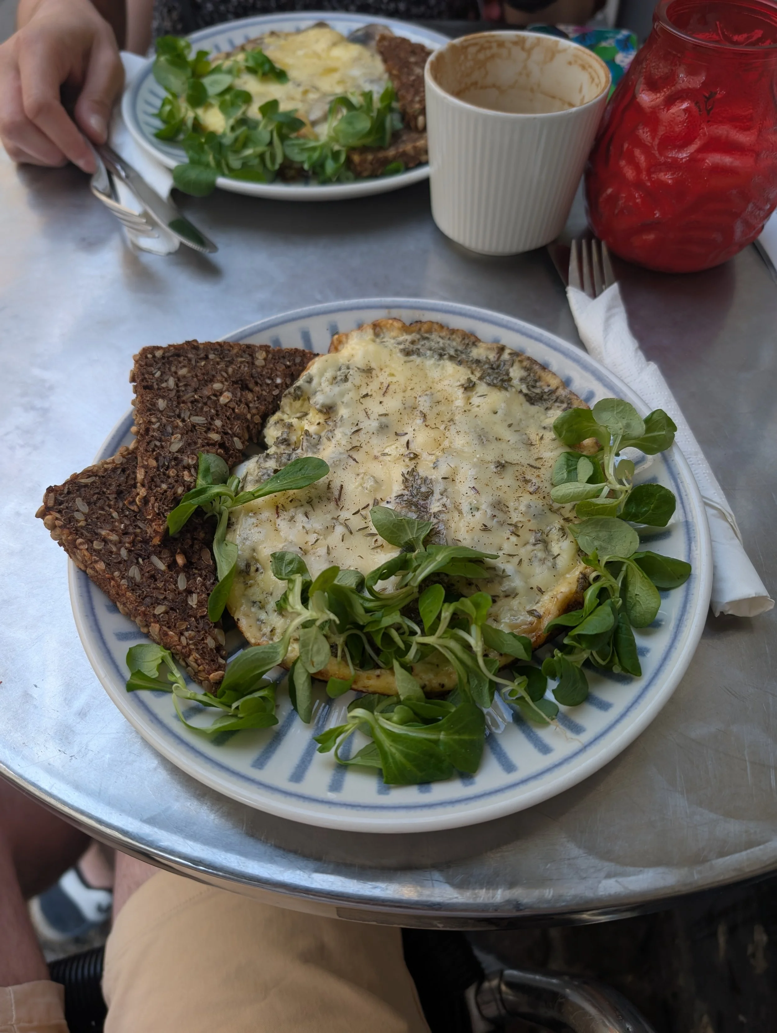 A plate of food with a slice of whole grain bread, a baked dish topped with melted cheese and herbs, garnished with fresh green herbs, on a table with a fork and napkin.