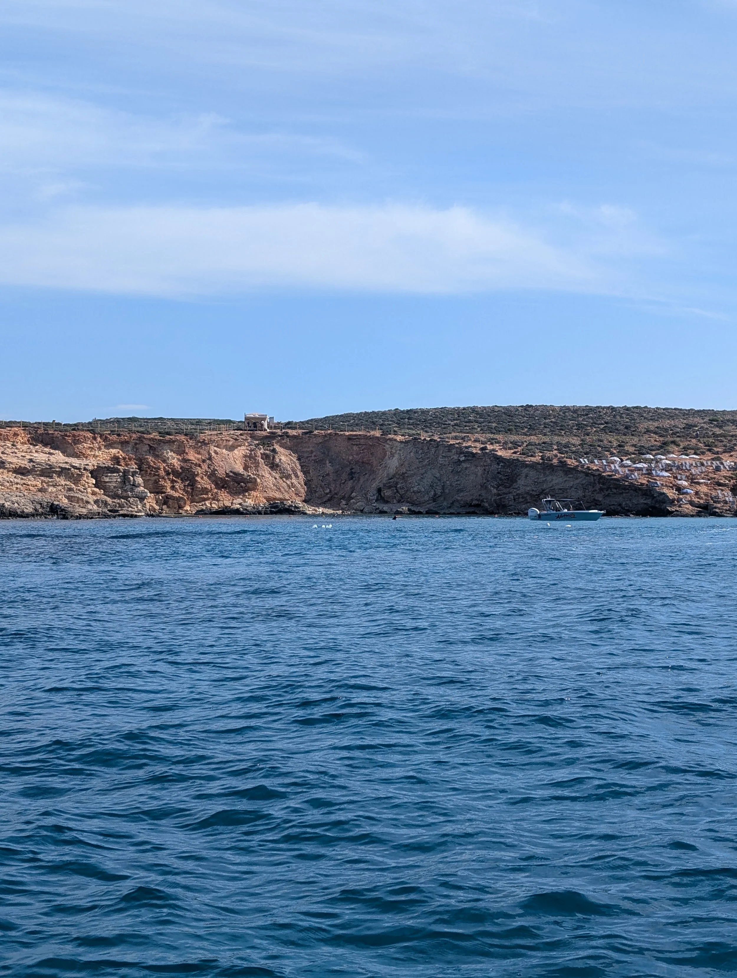 View of water in the foreground with rocky cliffs and a small white boat in the distance under a partly cloudy sky.
