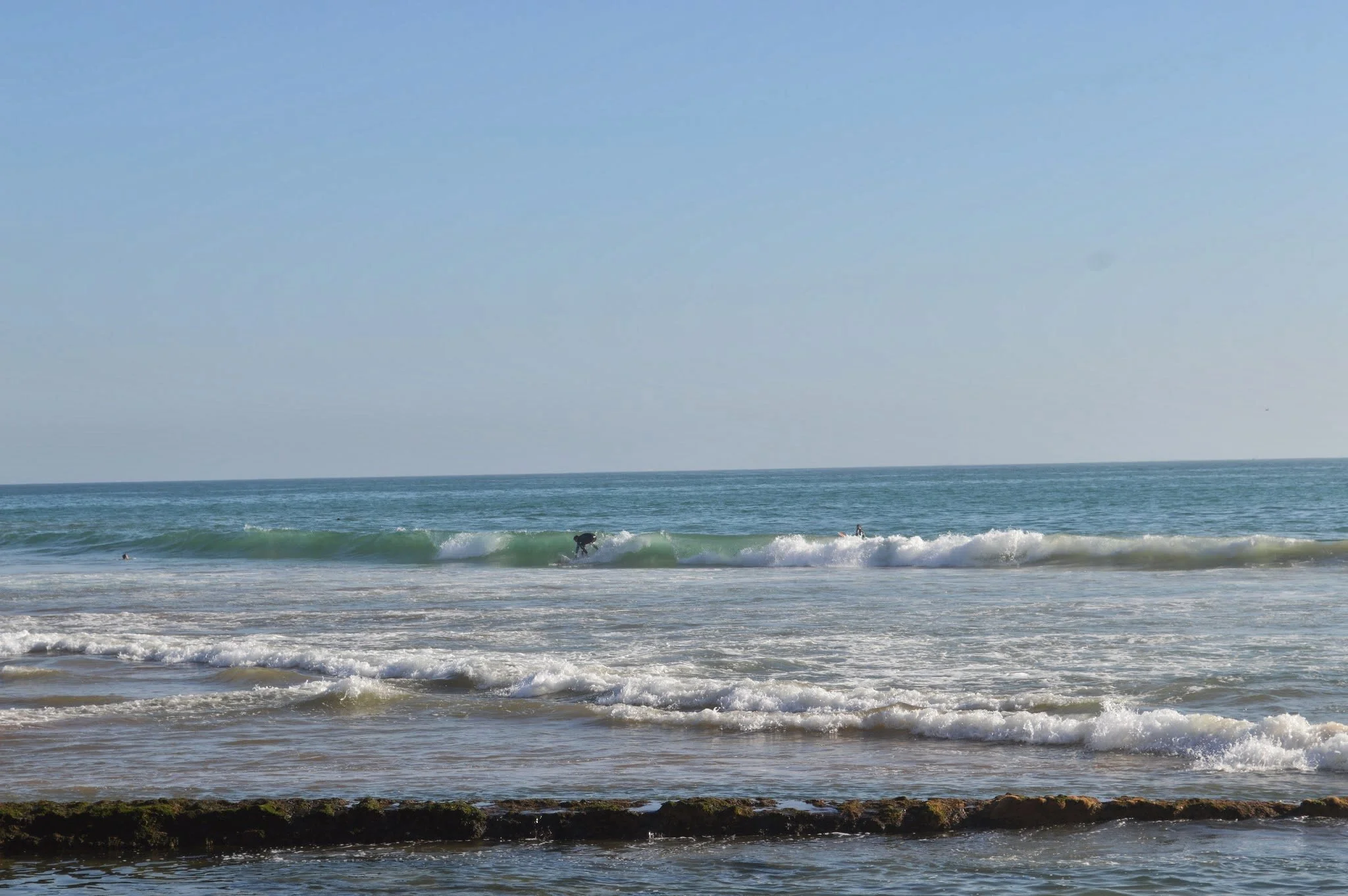A beach scene with blue sky, ocean waves, and two surfers in the water riding a wave.
