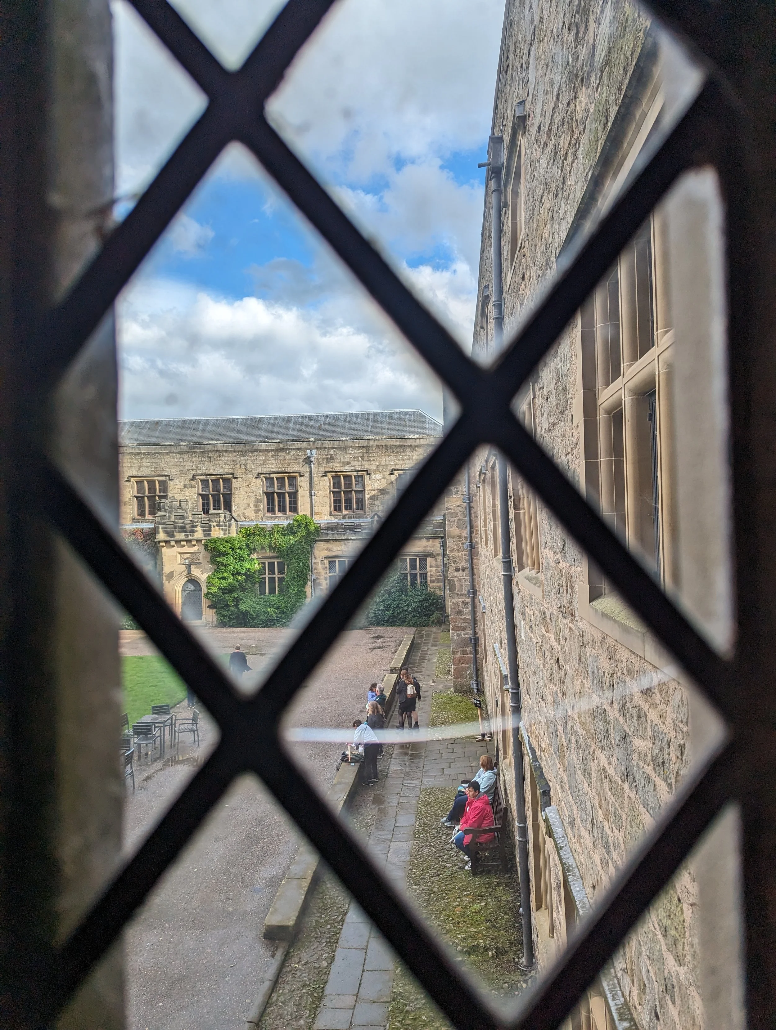 View through a metal lattice window showing a stone building with several windows, people sitting on benches and standing outside, some trees, and a blue sky with clouds.