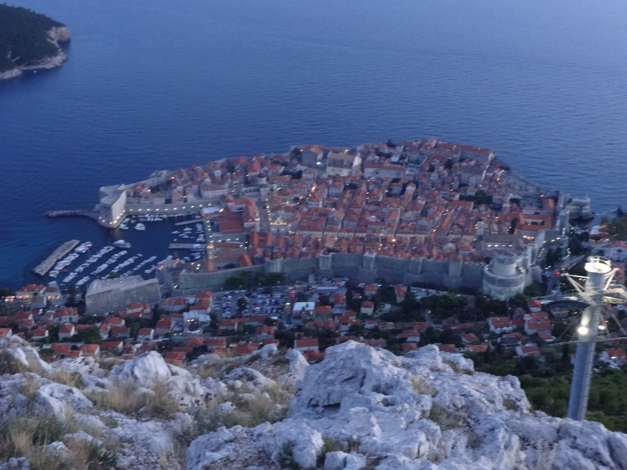 Aerial view of a coastal town with a historic fortress, red-tiled roofs, a marina with boats, and the sea in the background.