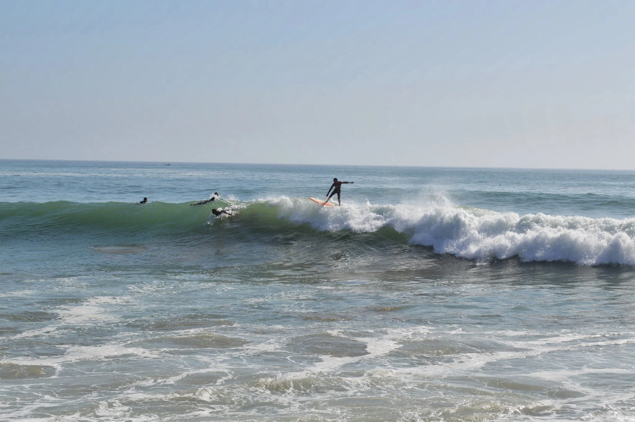 A person surfing on a wave at the beach with other surfers in the water and a boat in the distance under a clear sky.
