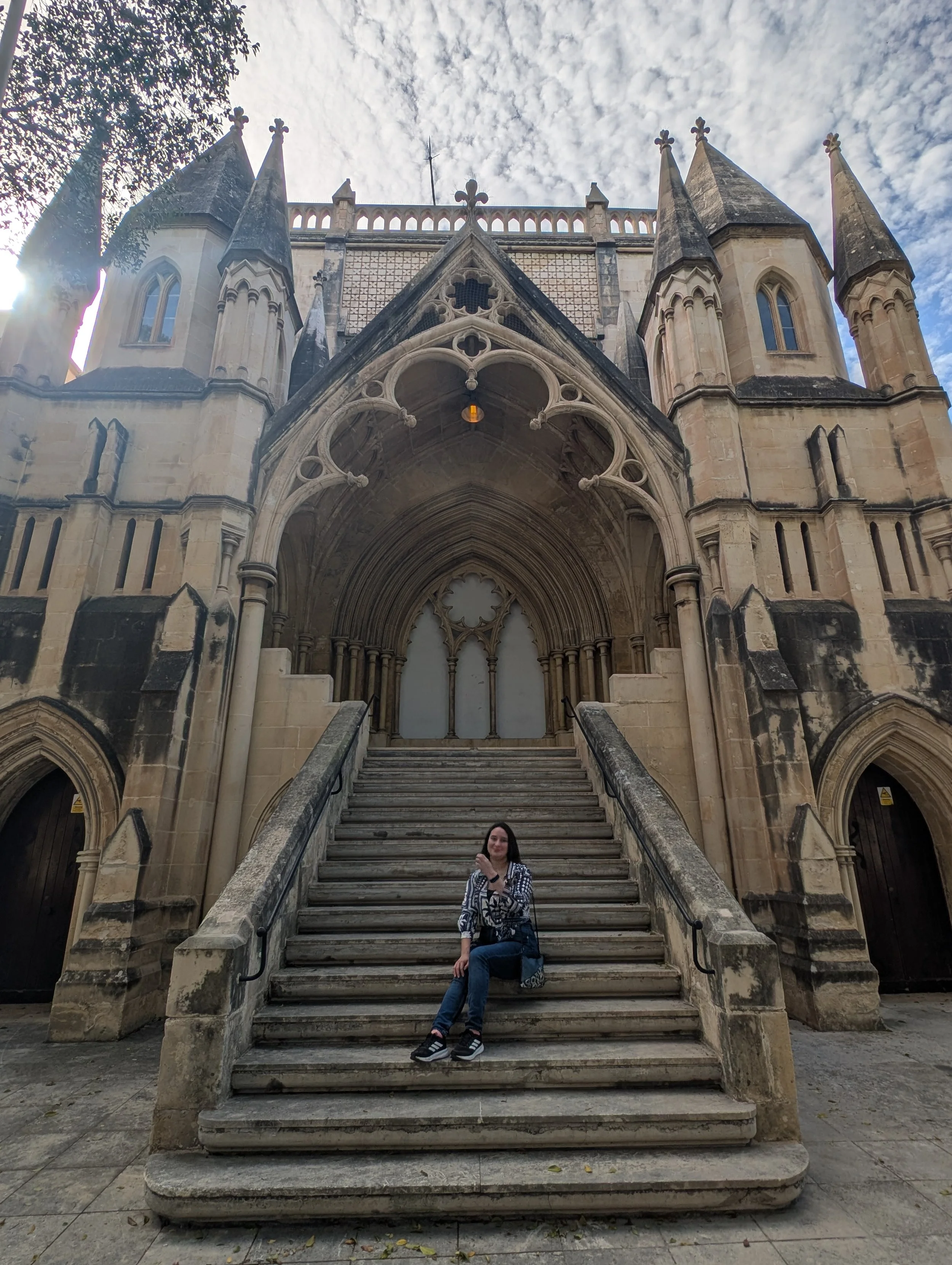A woman standing on the stairs of a historic Gothic-style church with pointed arches, decorative stonework, and tall spires, under a cloudy sky.