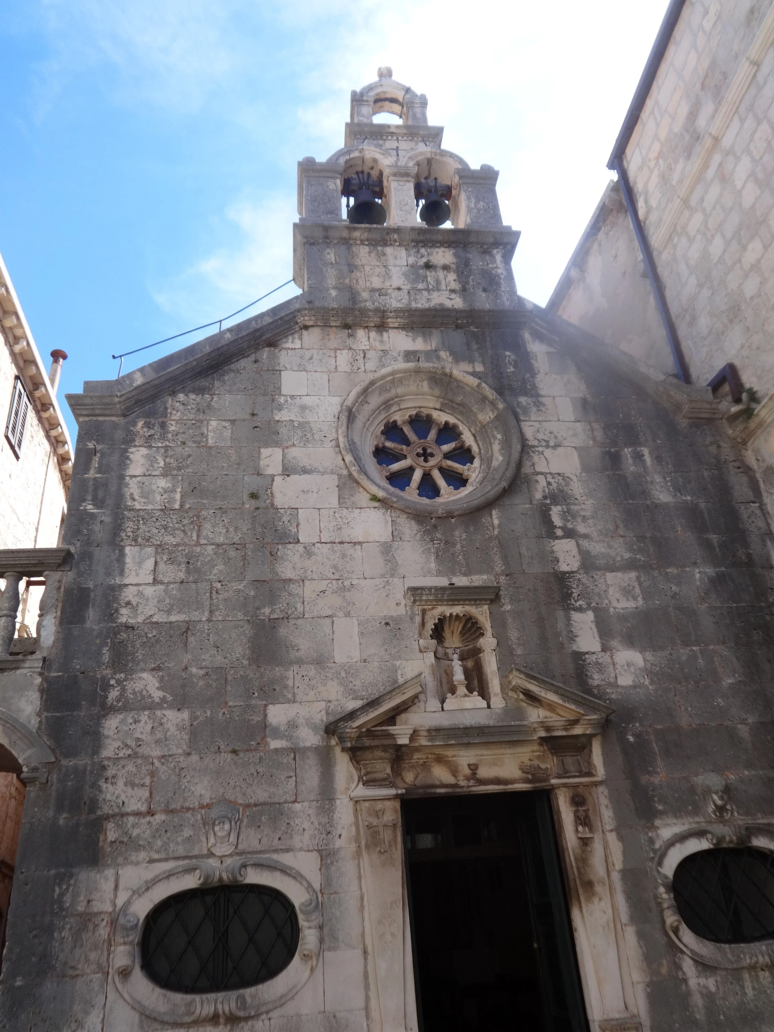 A historic stone church façade with circular windows, a small balcony with decorative columns, and a bell tower against a partly cloudy blue sky.