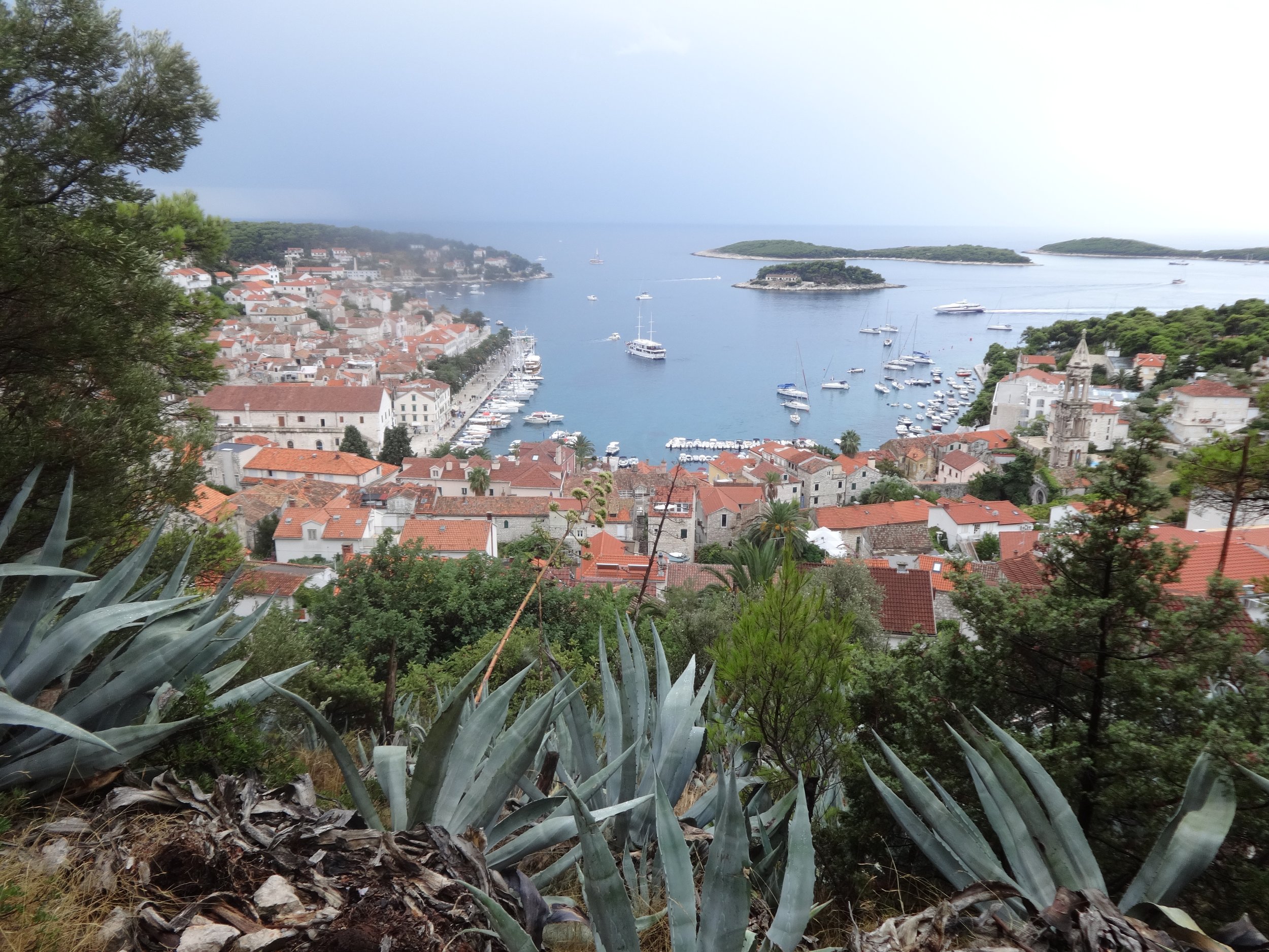 View of a coastal town with white buildings and red roofs along the shoreline, boats in the harbor, small islands in the water, and trees in the foreground.