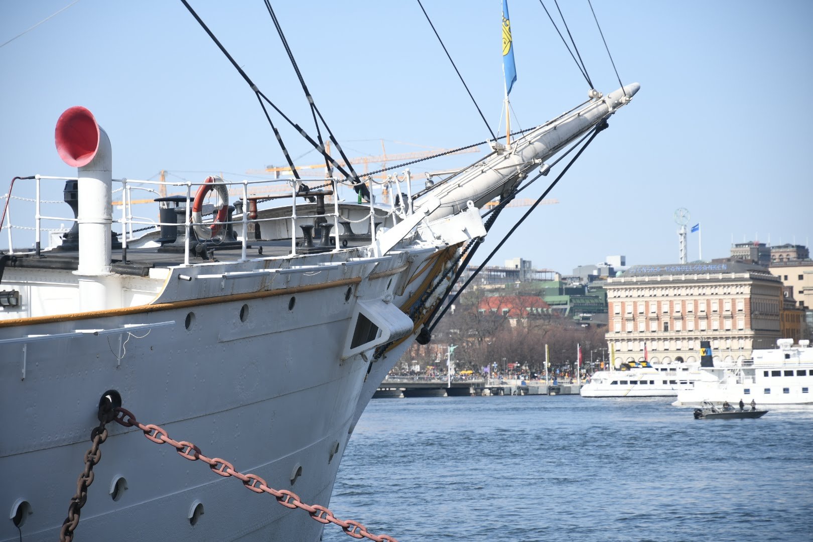 Close-up of a large white ship docked at a port, showing the bow, rigging, and part of the deck with a city skyline in the background.
