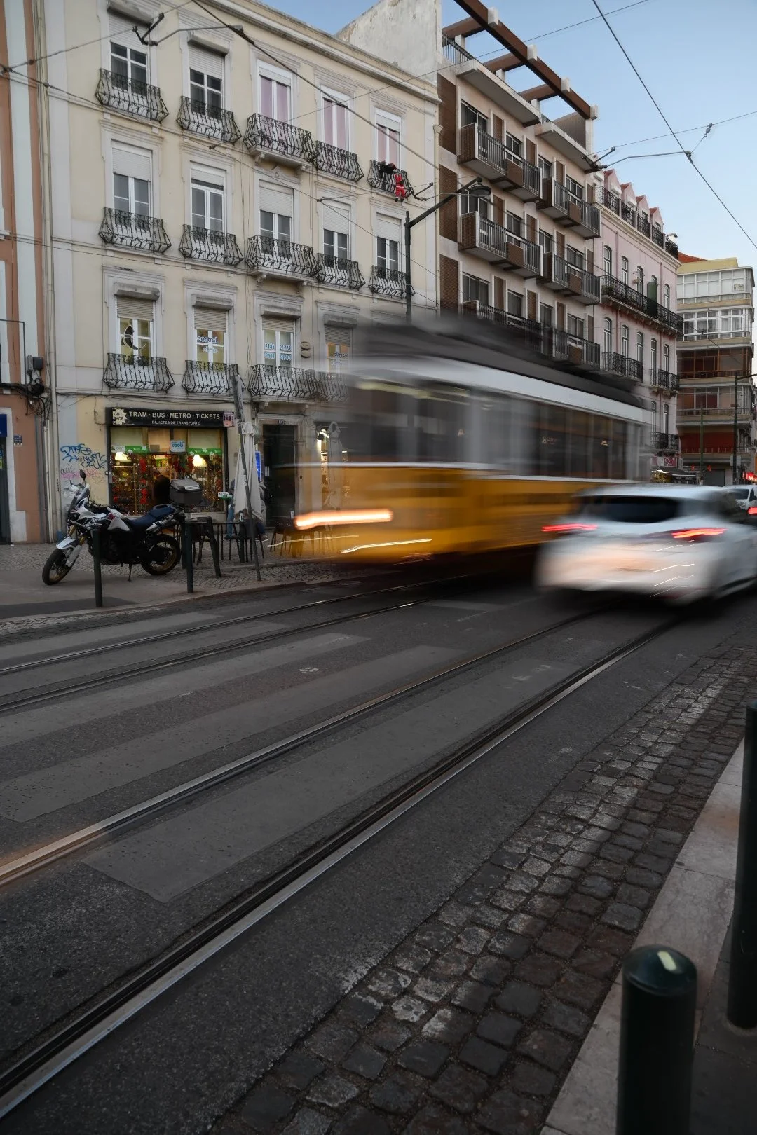 Blurred yellow tram moving along tracks on a city street, with a motorcycle and a white car passing by. Buildings with balconies and a store with a sign for tickets and transportation are visible in the background.