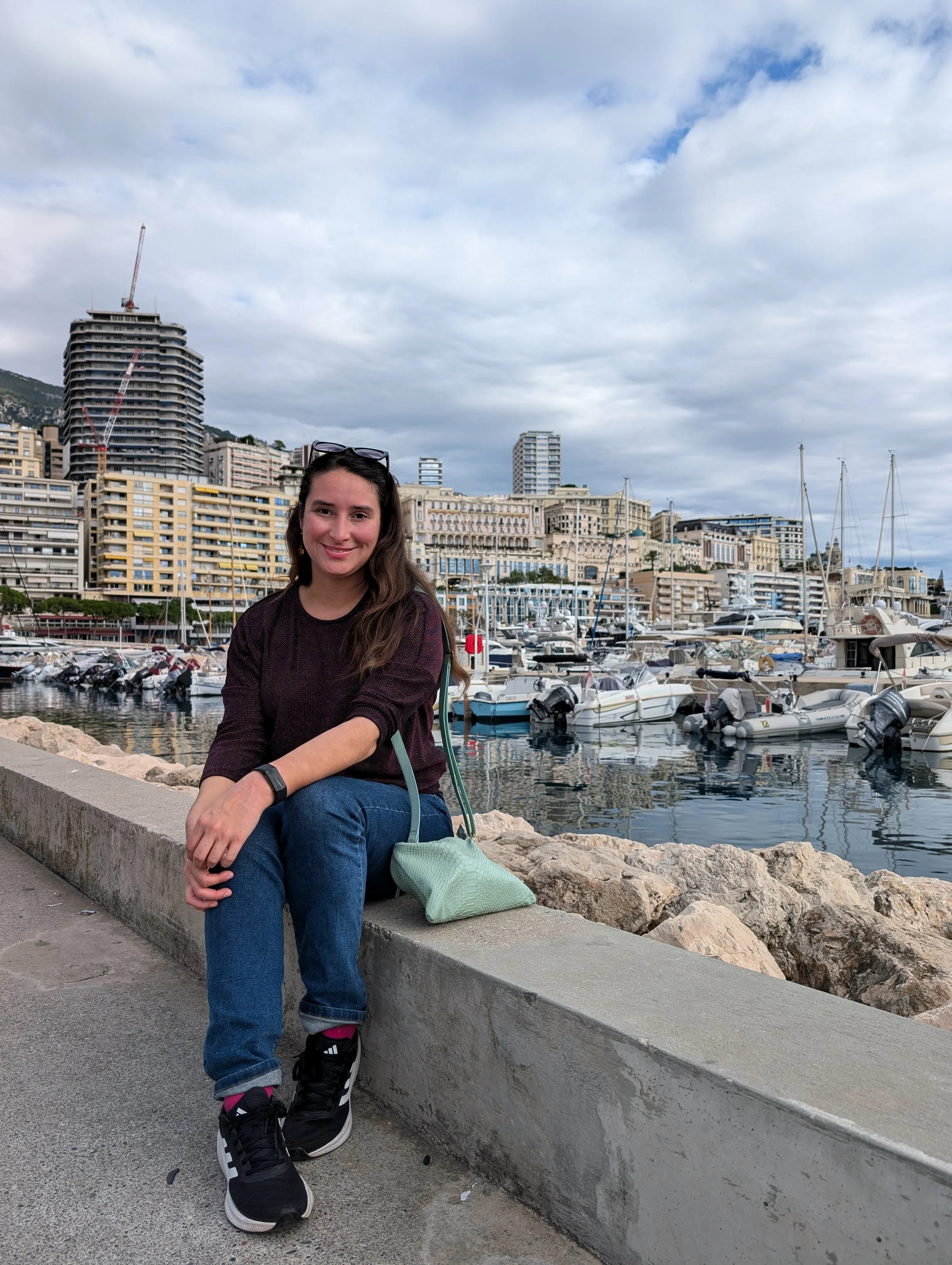A smiling woman with long brown hair, wearing a dark sweater and jeans, seated on a concrete ledge near a marina with boats, high-rise buildings, and a cloudy sky in the background.