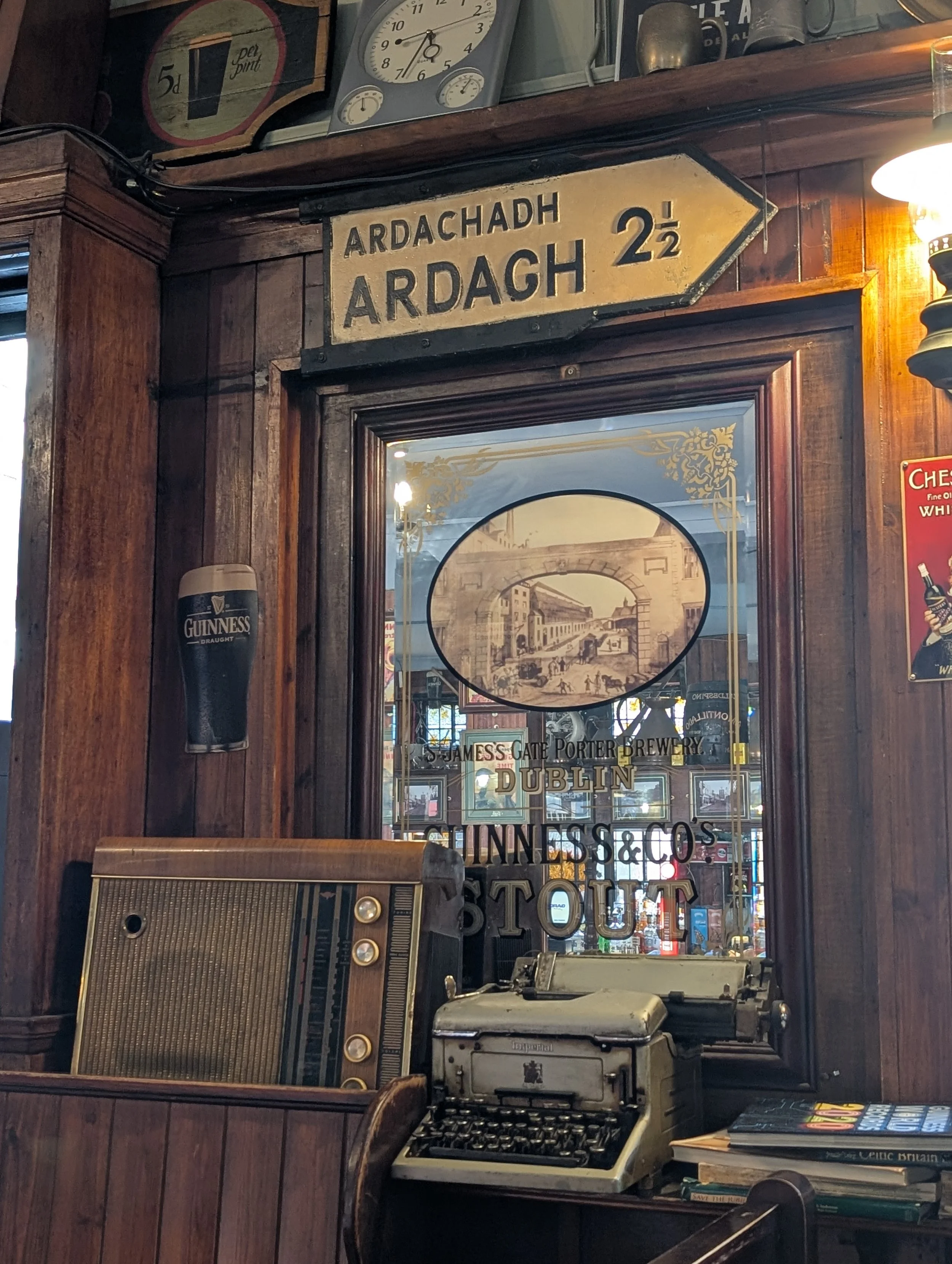 Vintage bar scene with wooden paneling, a sign pointing to Ardach 2 1/2 miles, a Guinness beer poster, a mirror reflecting a street scene, an old typewriter, and books.