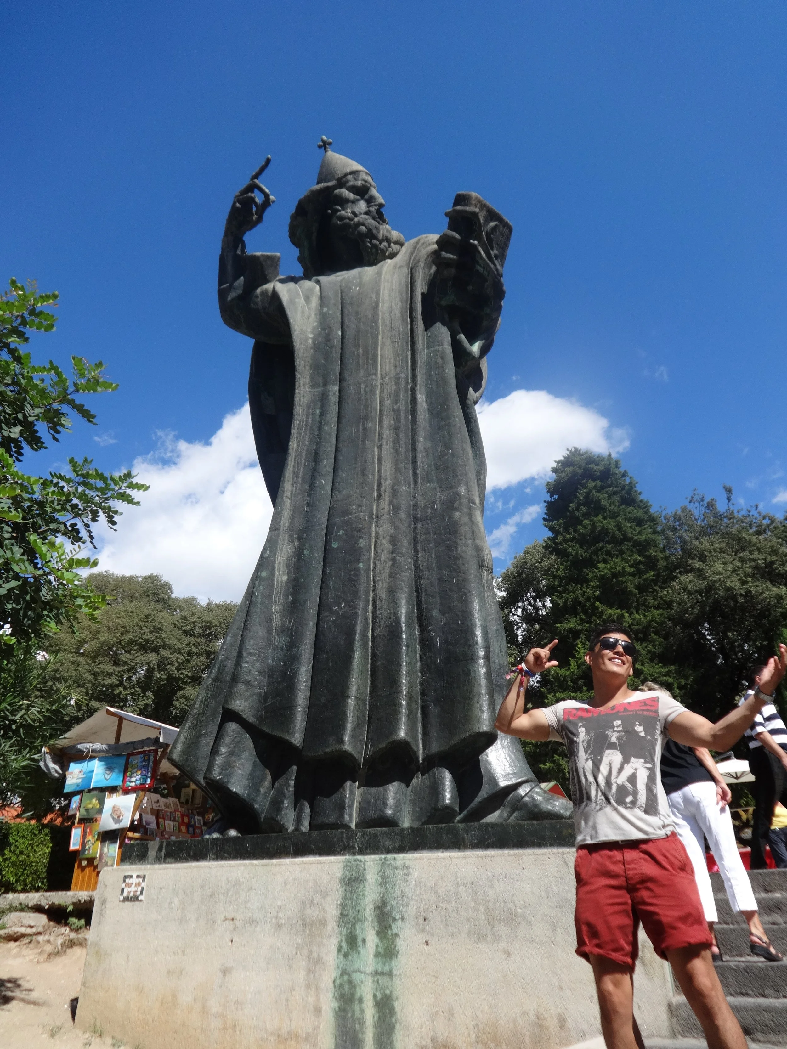 A large stone statue of a bearded man with a pointed hat, holding a book, with a person in sunglasses and a graphic t-shirt smiling and pointing at the statue in an outdoor park setting.