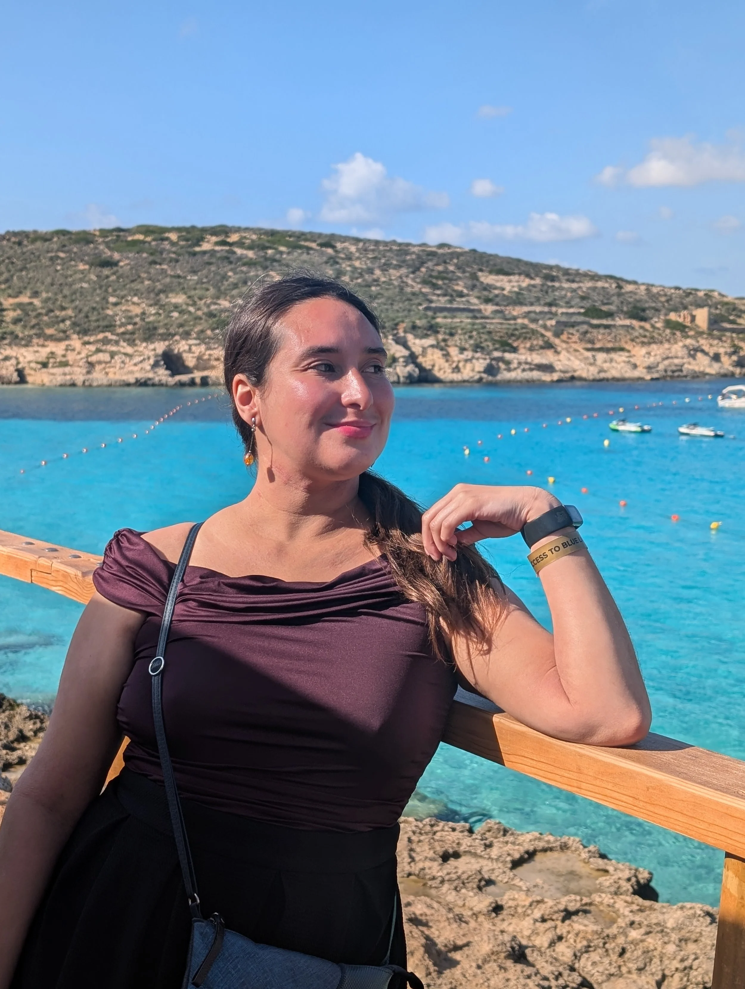 A woman with dark hair, wearing earrings and a maroon shirt, sitting on a wooden rail near a body of water with boats and rocky shoreline, with a hill and blue sky in the background.