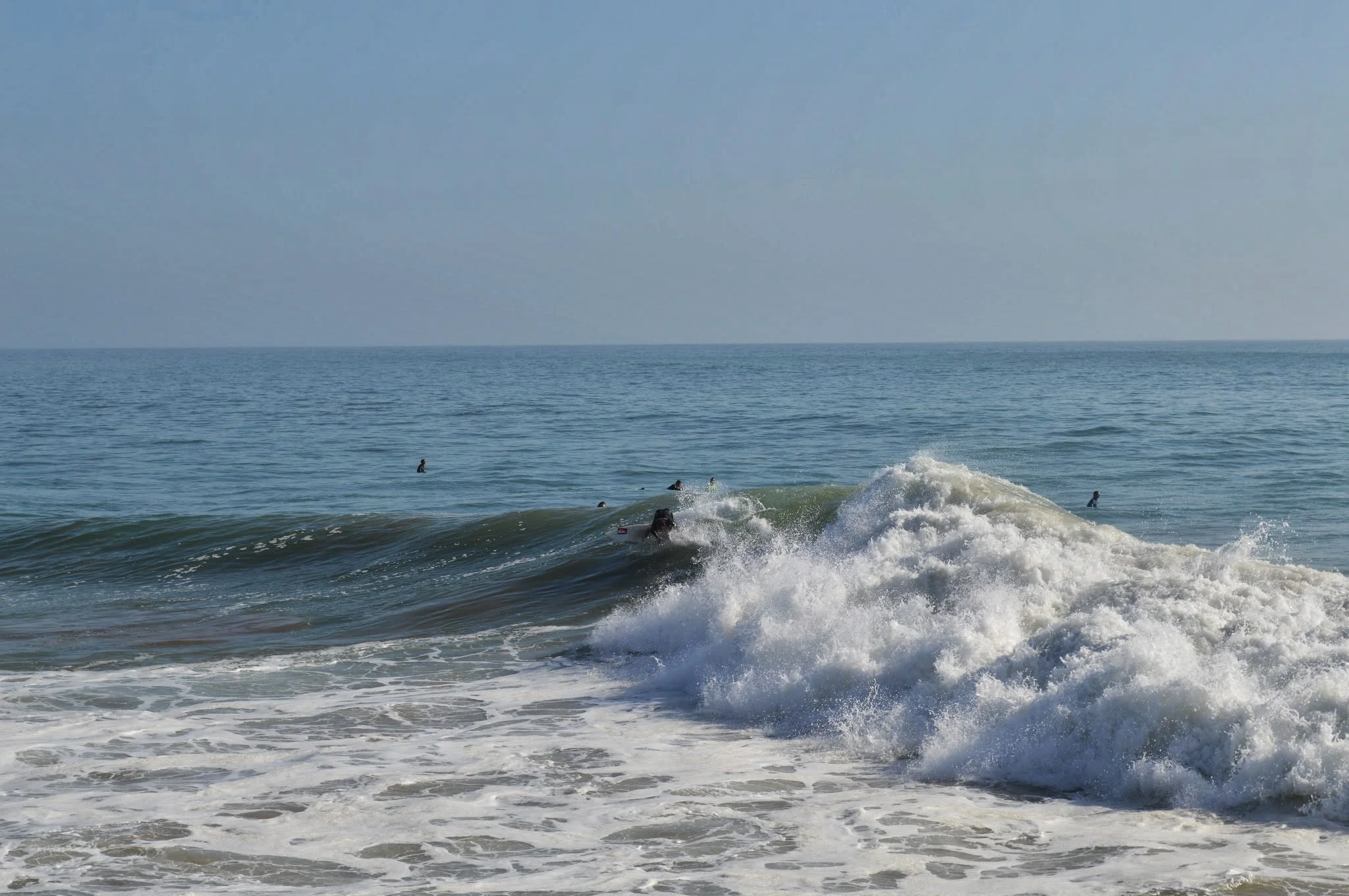 Beach scene with blue ocean waves, a few people swimming or surfing in the water, and a light blue sky.