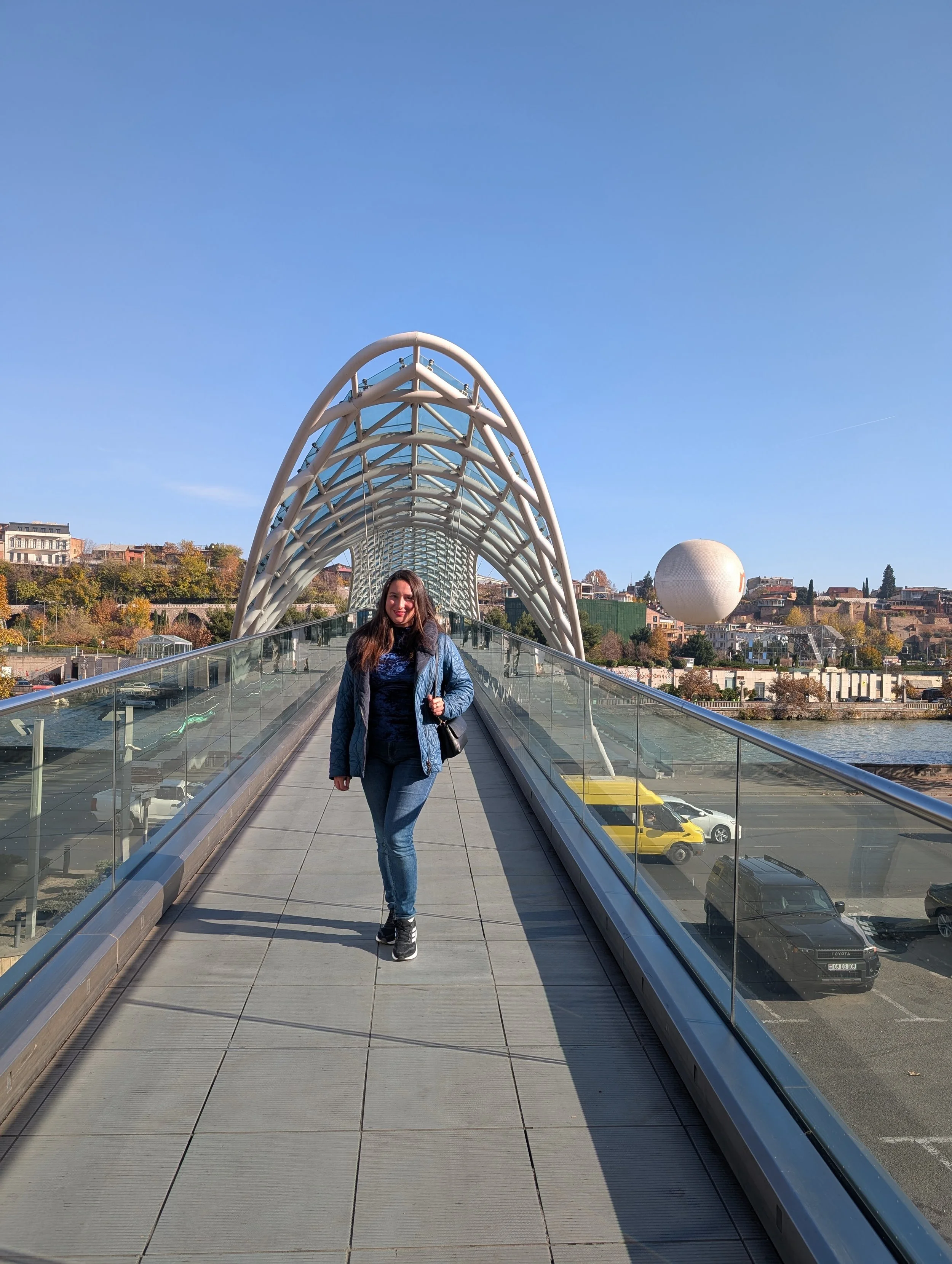 A young woman walking on a modern glass bridge with a metallic arch design in a cityscape, with residential and commercial buildings, a spherical structure, and cars below under a clear blue sky.