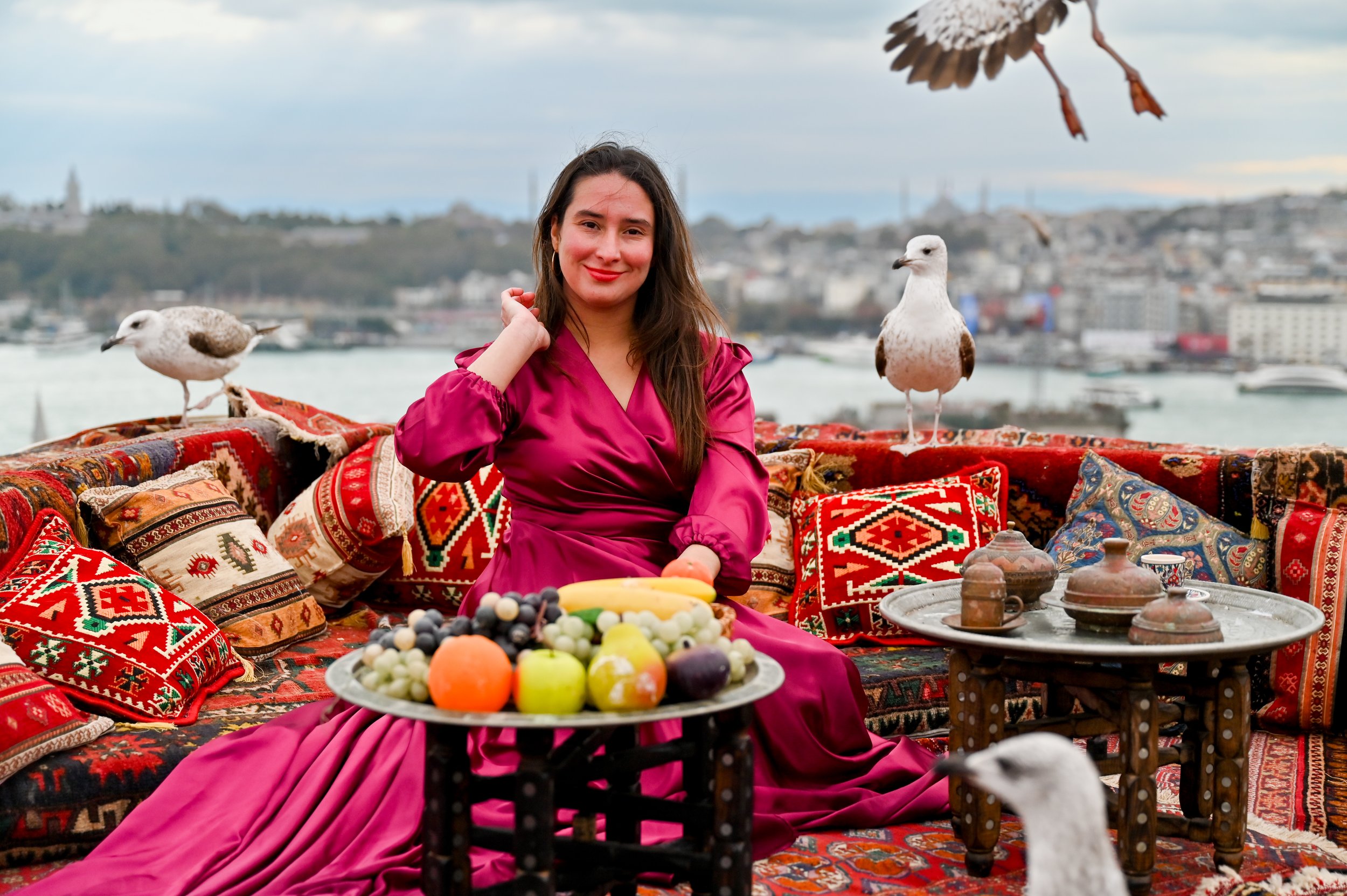 A woman in a vibrant pink dress sitting on a traditional patterned rug with Persian pillows, surrounded by seagulls, with a city waterfront and boats in the background, and various fruits on a table in front.