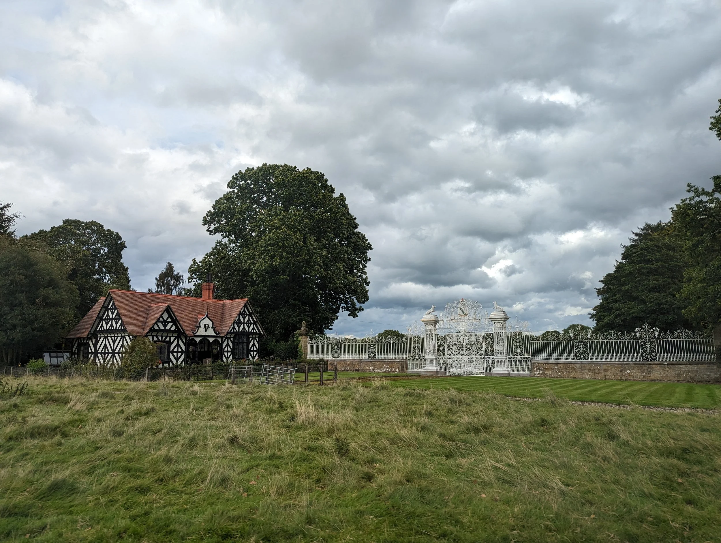 A house with a black and white Tudor-style facade, red roof, and a large green tree in front. There is a white ornate gate in the background, and a cloudy sky overhead.