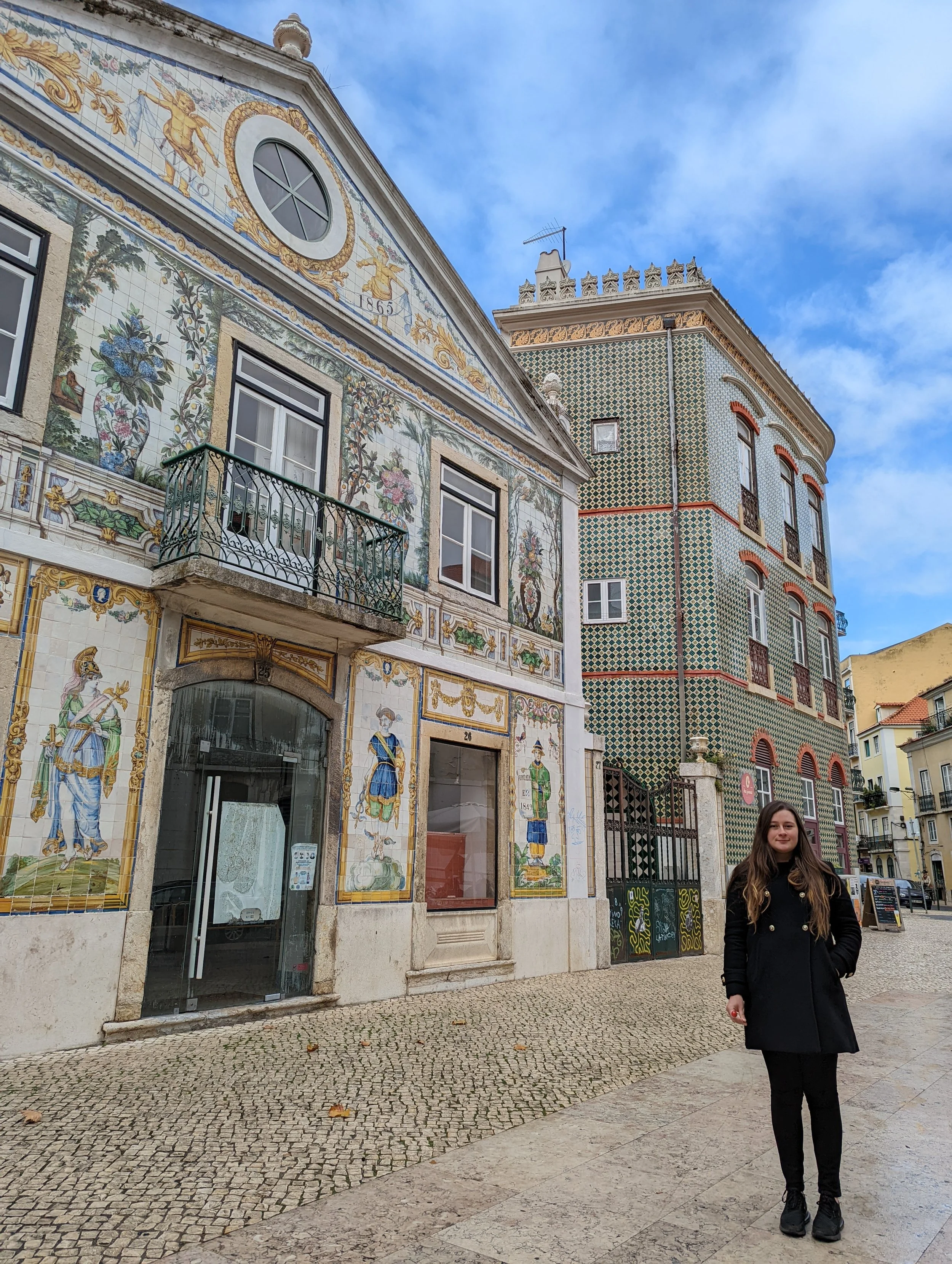 A woman with long brown hair wearing a black coat and black pants standing on a cobblestone street in front of a decorated building with colorful ceramic tiles and novel artwork, with a blue sky overhead.