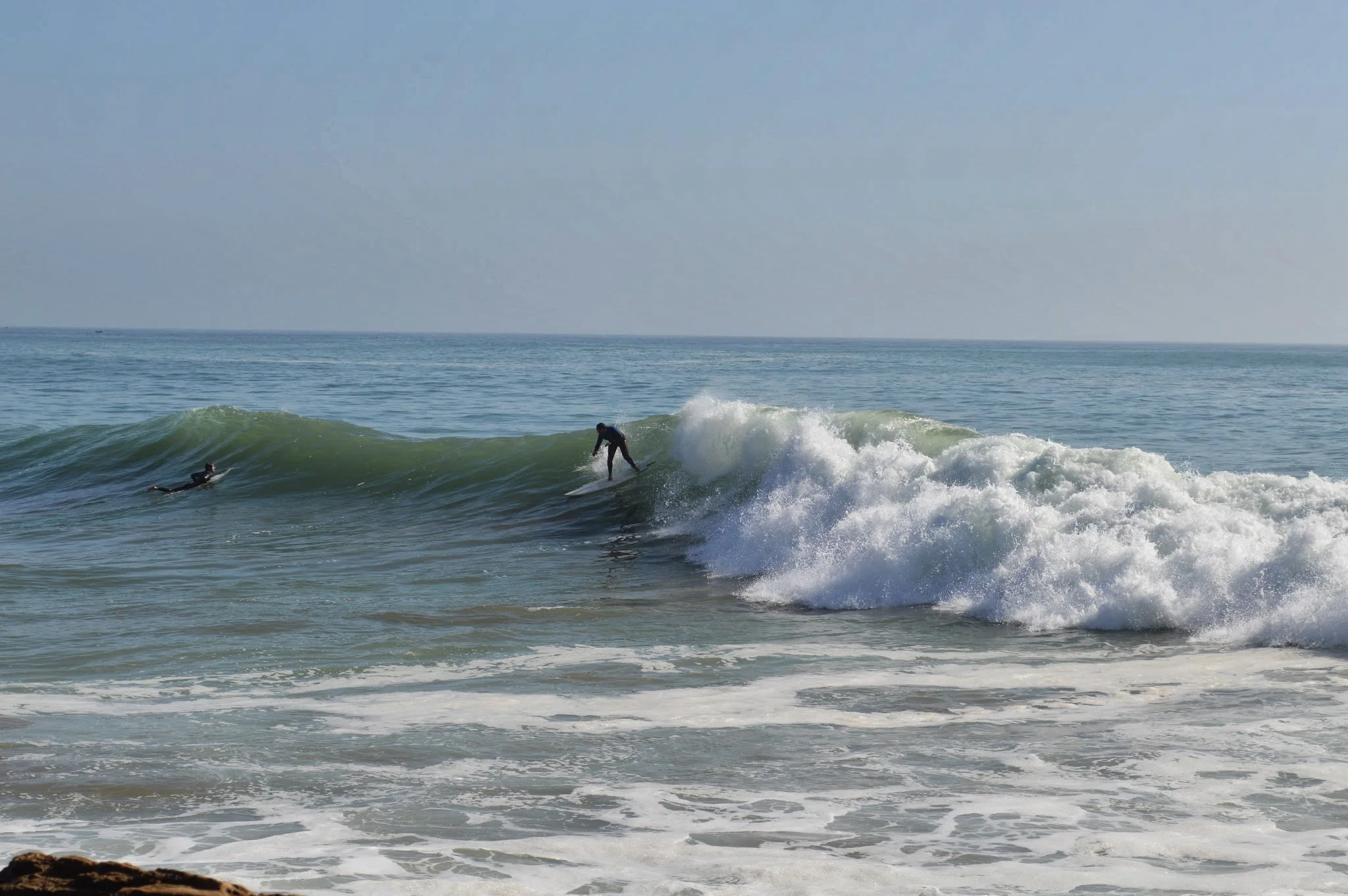 Two surfers riding or preparing to ride ocean waves near the shoreline with a clear sky and calm sea in the background.