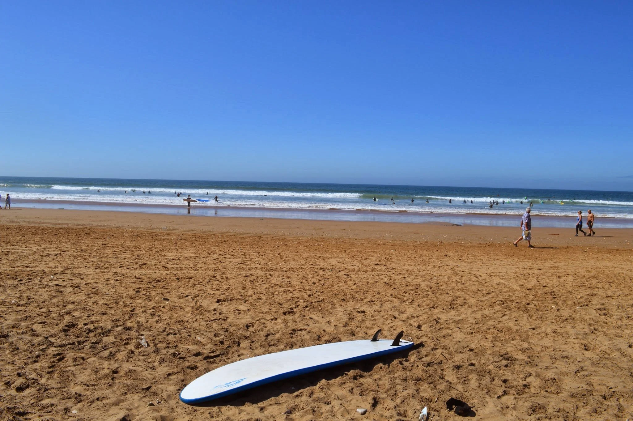 View of a sandy beach with a surfboard in the foreground, people swimming and surfing in the ocean, and some walking along the shoreline under a clear blue sky.