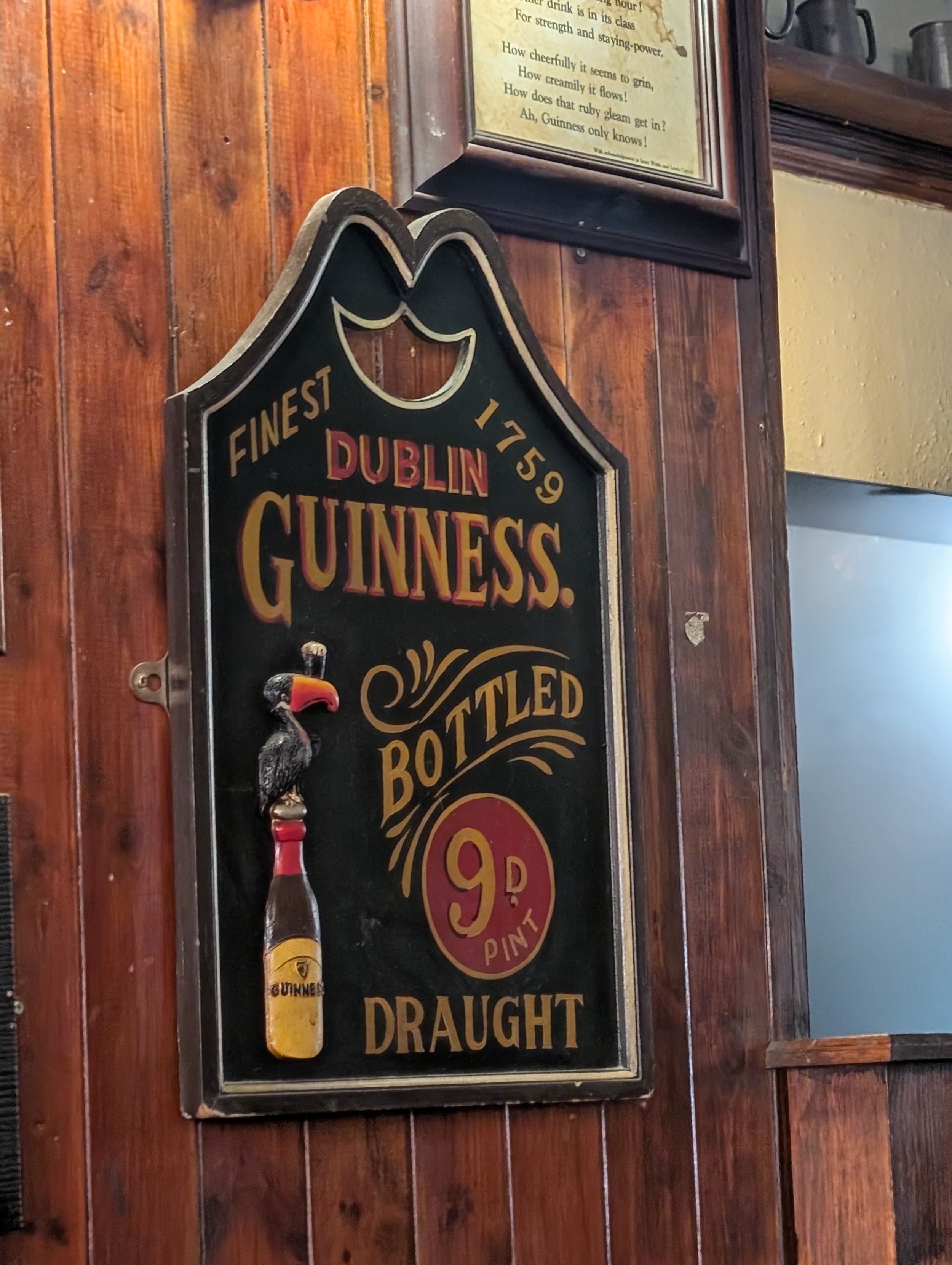 A black sign with gold and red text displaying prices for Guinness draught beer in a bar with wooden walls, decorated with a decorative Guinness bottle opener shaped like a pint of beer and a small figurine of a bird.