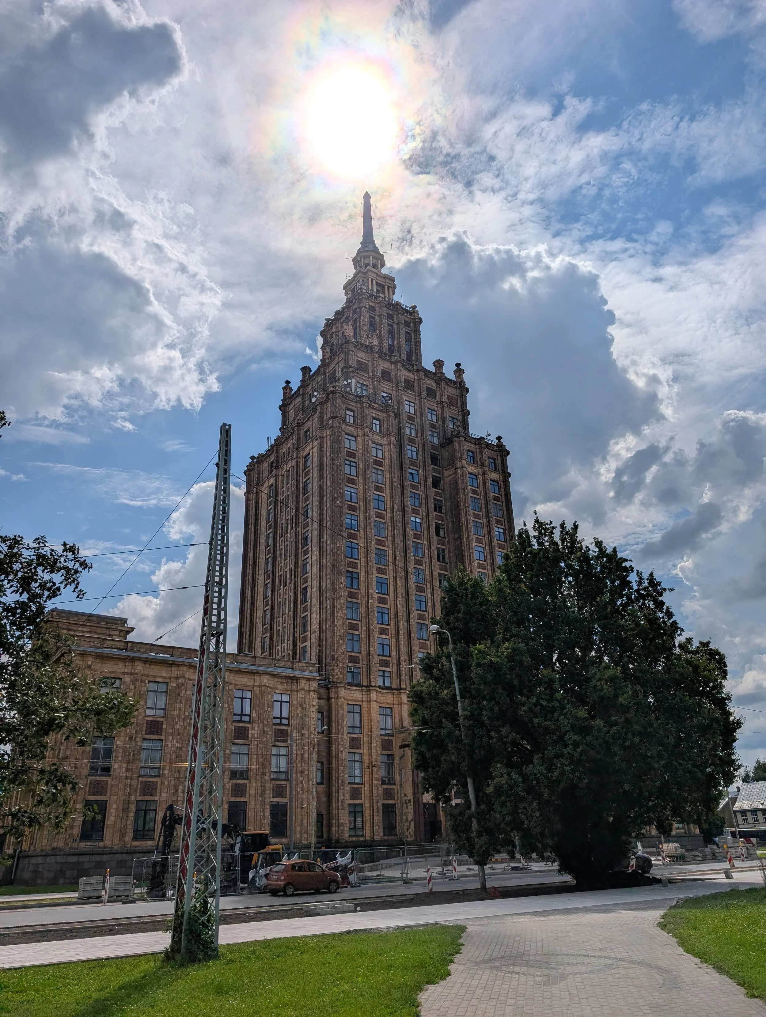 A tall, historic building with a spire, parked cars on the street, trees, and a cloudy sky with the sun partially covered by clouds.