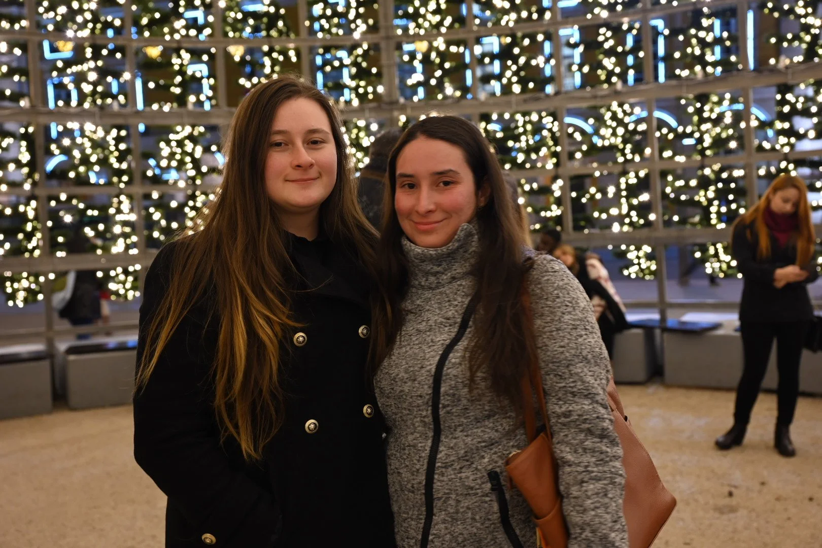 Two women standing in front of a decorated Christmas tree with lights, indoors, smiling at the camera.