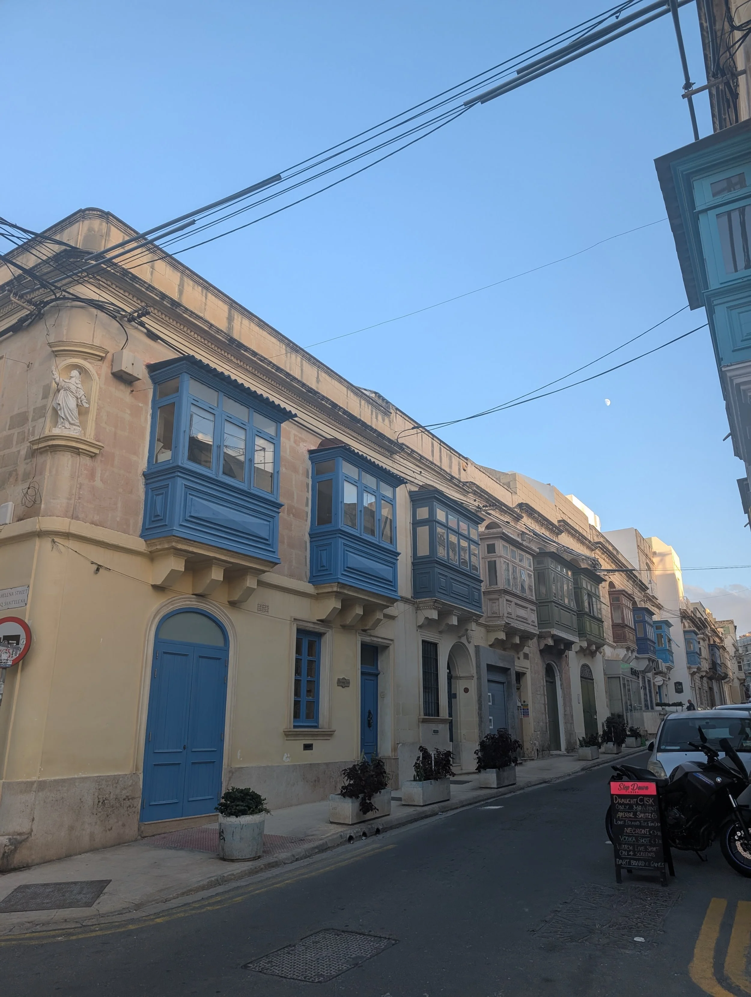 Street view of old buildings with blue balconies and shutters, potted plants along the sidewalk, utility wires overhead, and a small chalkboard sign advertising drinks and snacks, with a clear blue sky and a visible moon.