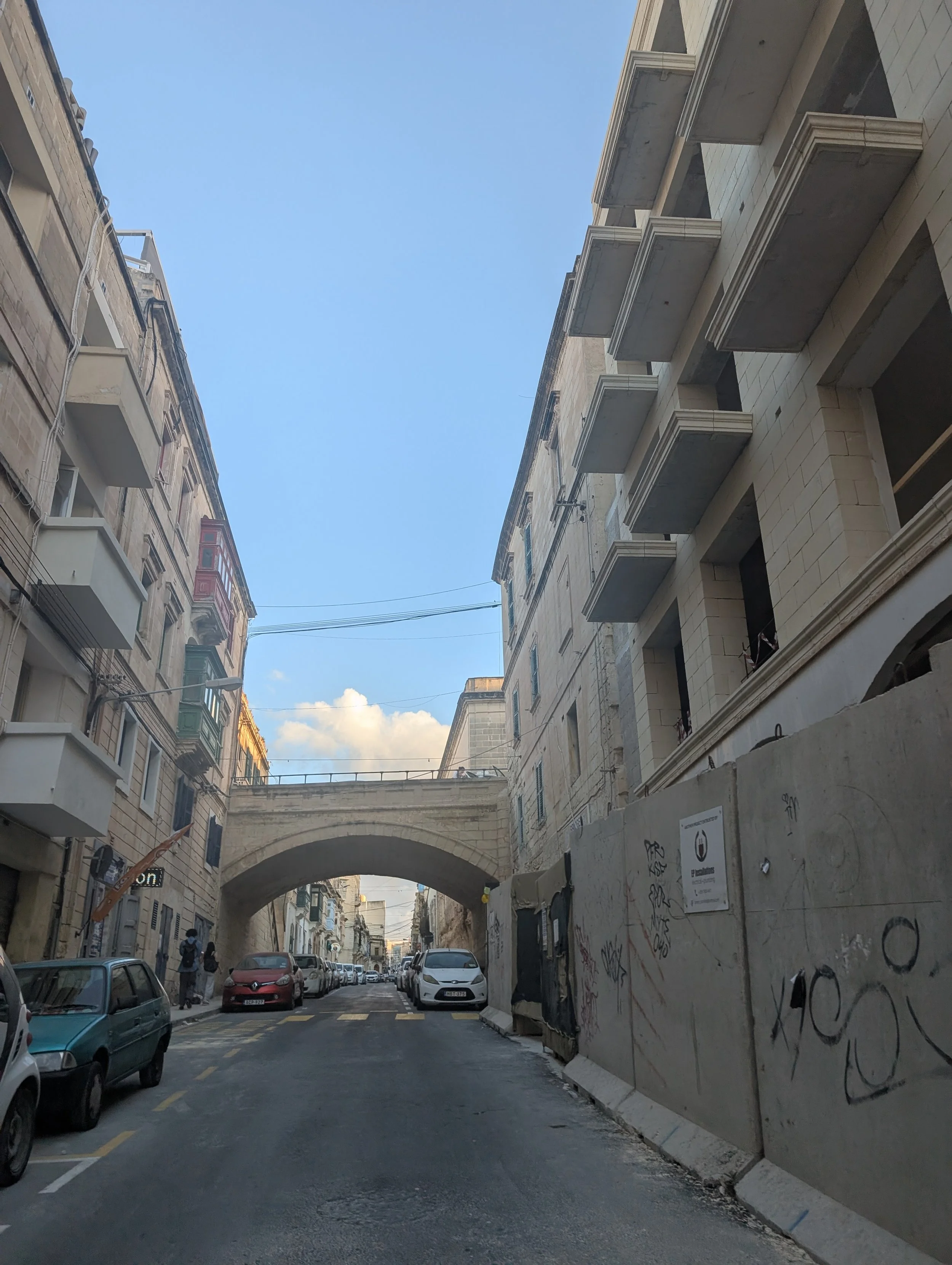 A narrow city street with cars parked along both sides, and a stone arch bridge connecting two buildings. The buildings are multi-story with balconies and graffiti on the walls, under a clear blue sky.