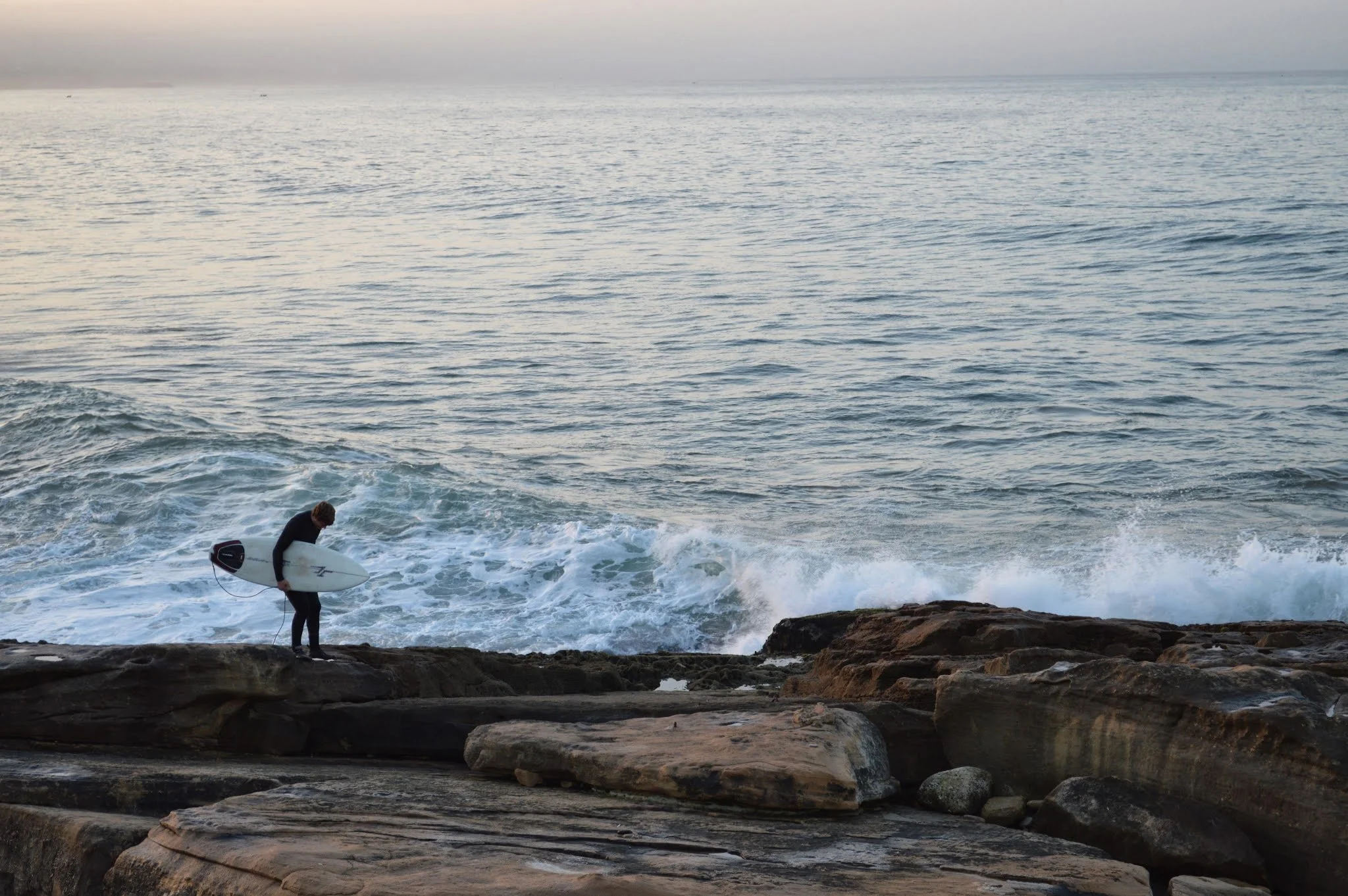 A person in a wetsuit stands on a rocky shoreline holding a surfboard, preparing to surf in the ocean near the rocks with gentle waves, under a cloudy sky.