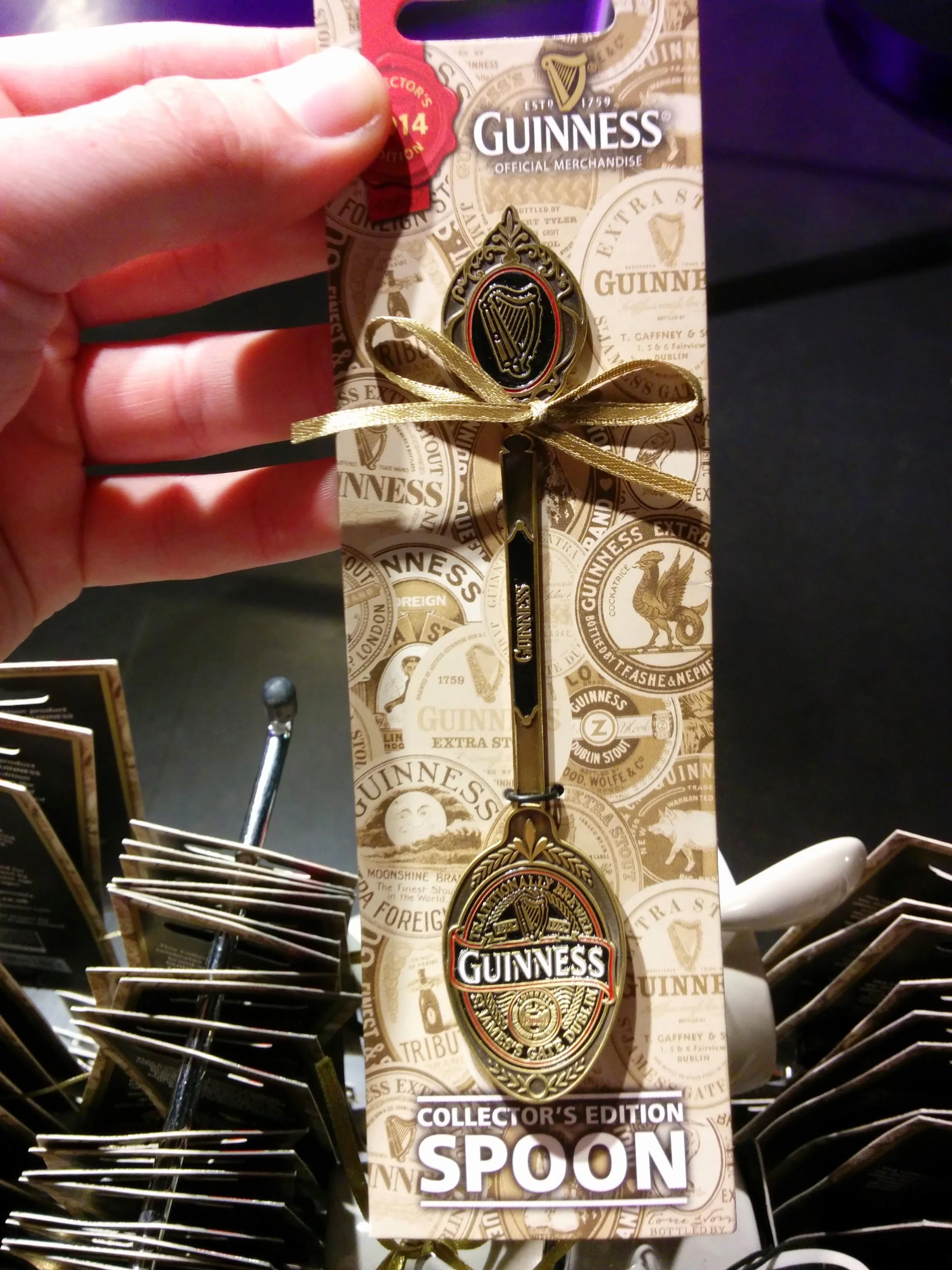 A collectible Guinness Irish pub spoon in its packaging, displayed on a store rack. The spoon features a Guinness logo and decorative design, with a gold-colored ribbon tied around it. The packaging has a beige background with various Guinness-themed