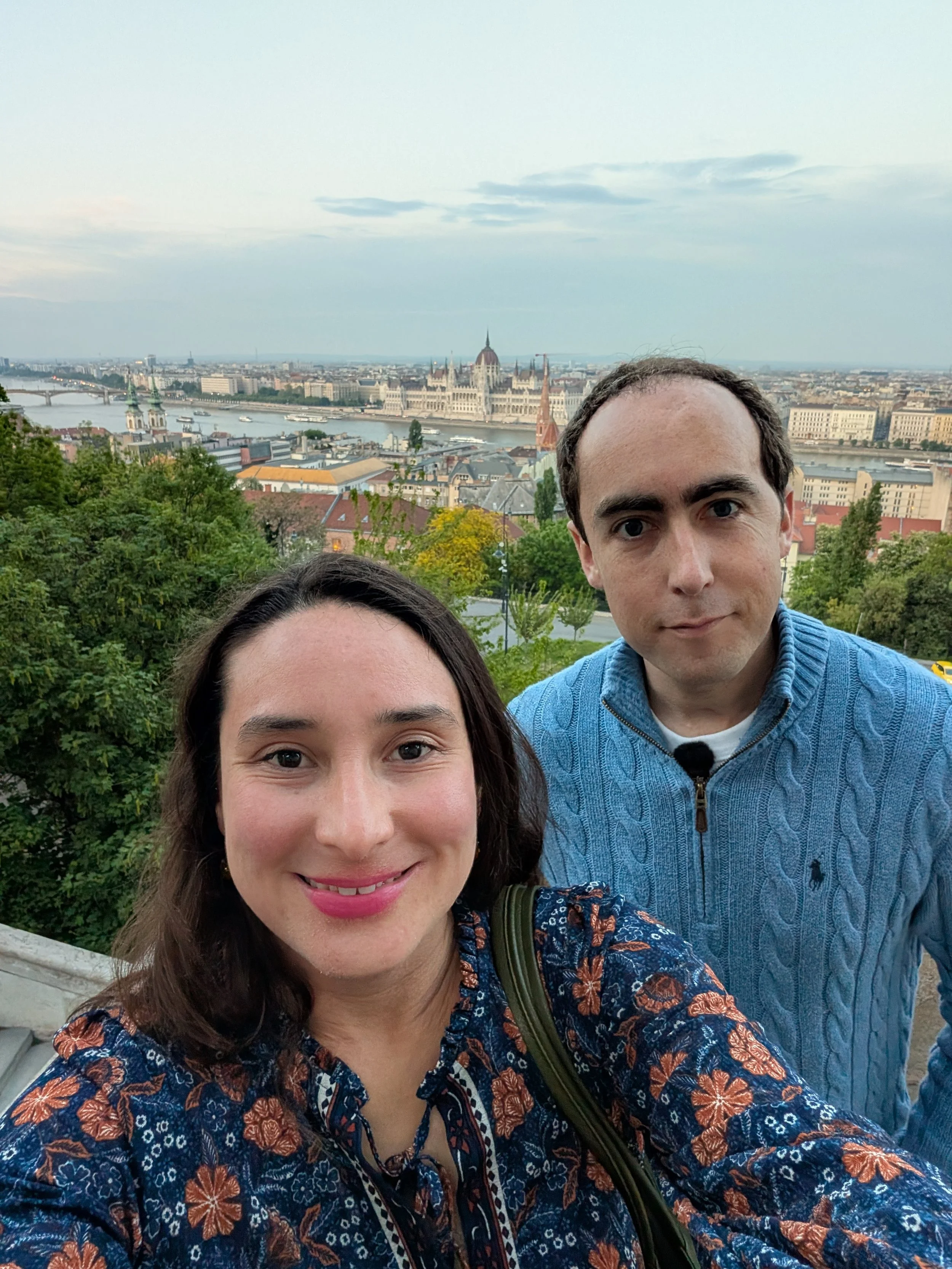 A man and woman taking a selfie outdoors with a view of the city and a river in the background. The woman has brown hair and is smiling, wearing a floral-patterned jacket. The man has dark hair, wearing a blue sweater, and has a neutral expression. T
