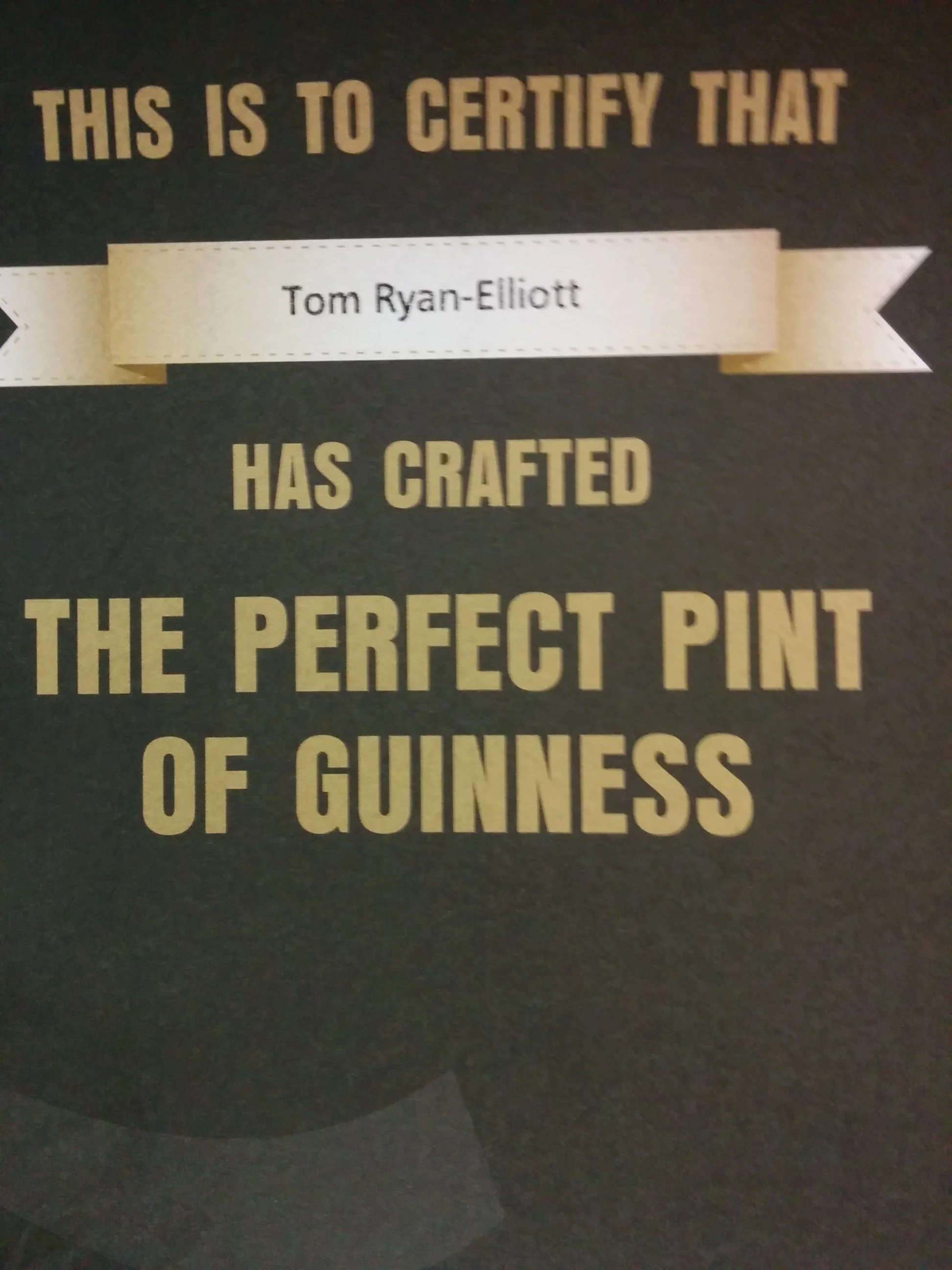 Part of a sign or plaque with the text 'This is to certify that Tom Ryan-Elliott has crafted the perfect pint of Guinness.' The text is printed on a black background with some white and gold-colored accents.
