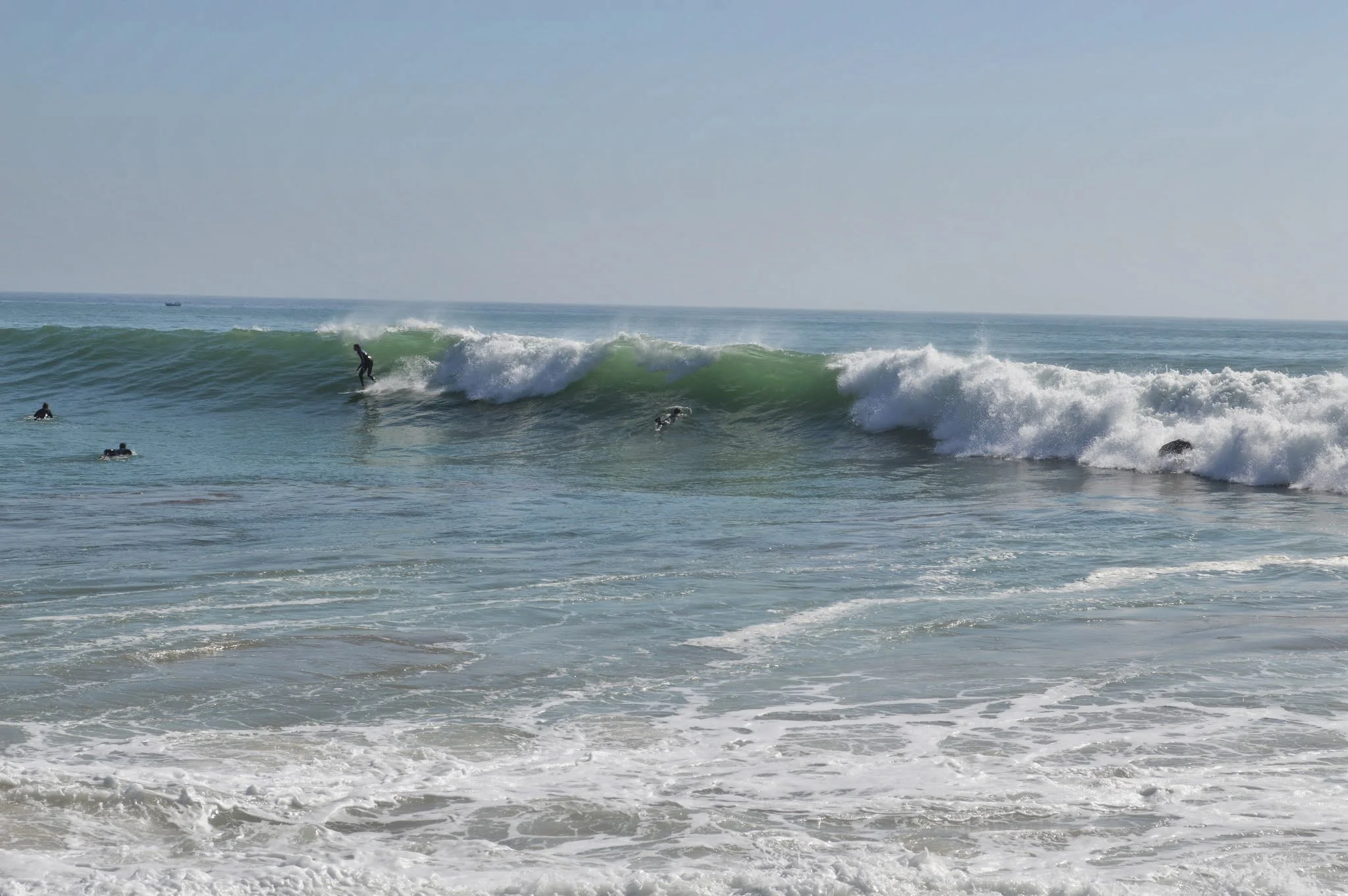 People surfing on waves in the ocean near the shoreline with a boat in the distance under a clear sky.