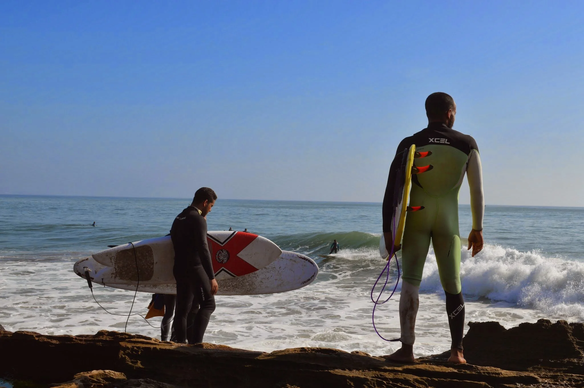 Two surfers preparing to enter the water on a sunny day at the beach, one standing with a surfboard and the other holding a surfboard, with the ocean and blue sky in the background.