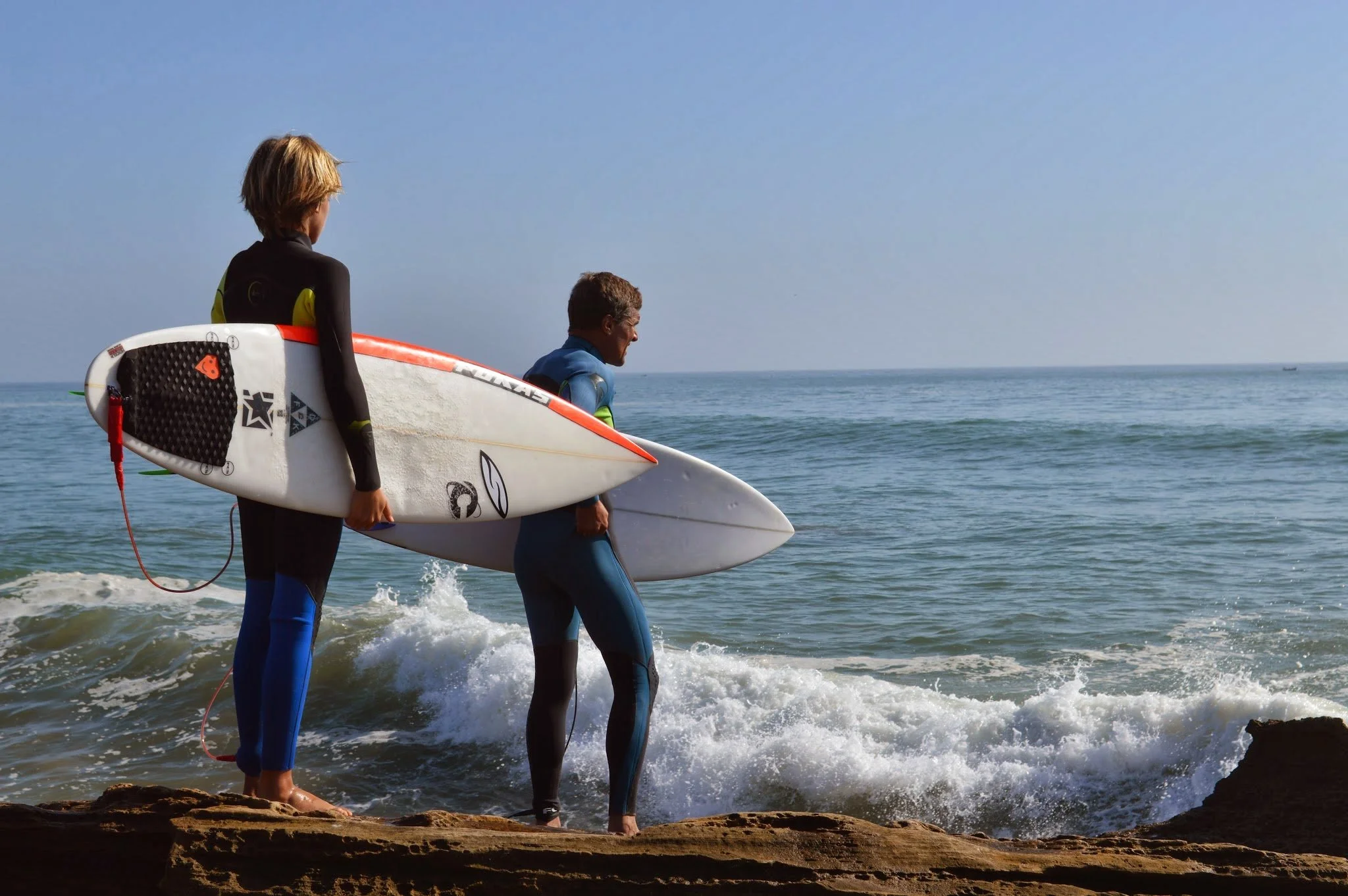 Two surfers in wetsuits walking along the rocky shoreline holding surfboards, with the ocean waves at their feet under a clear blue sky.