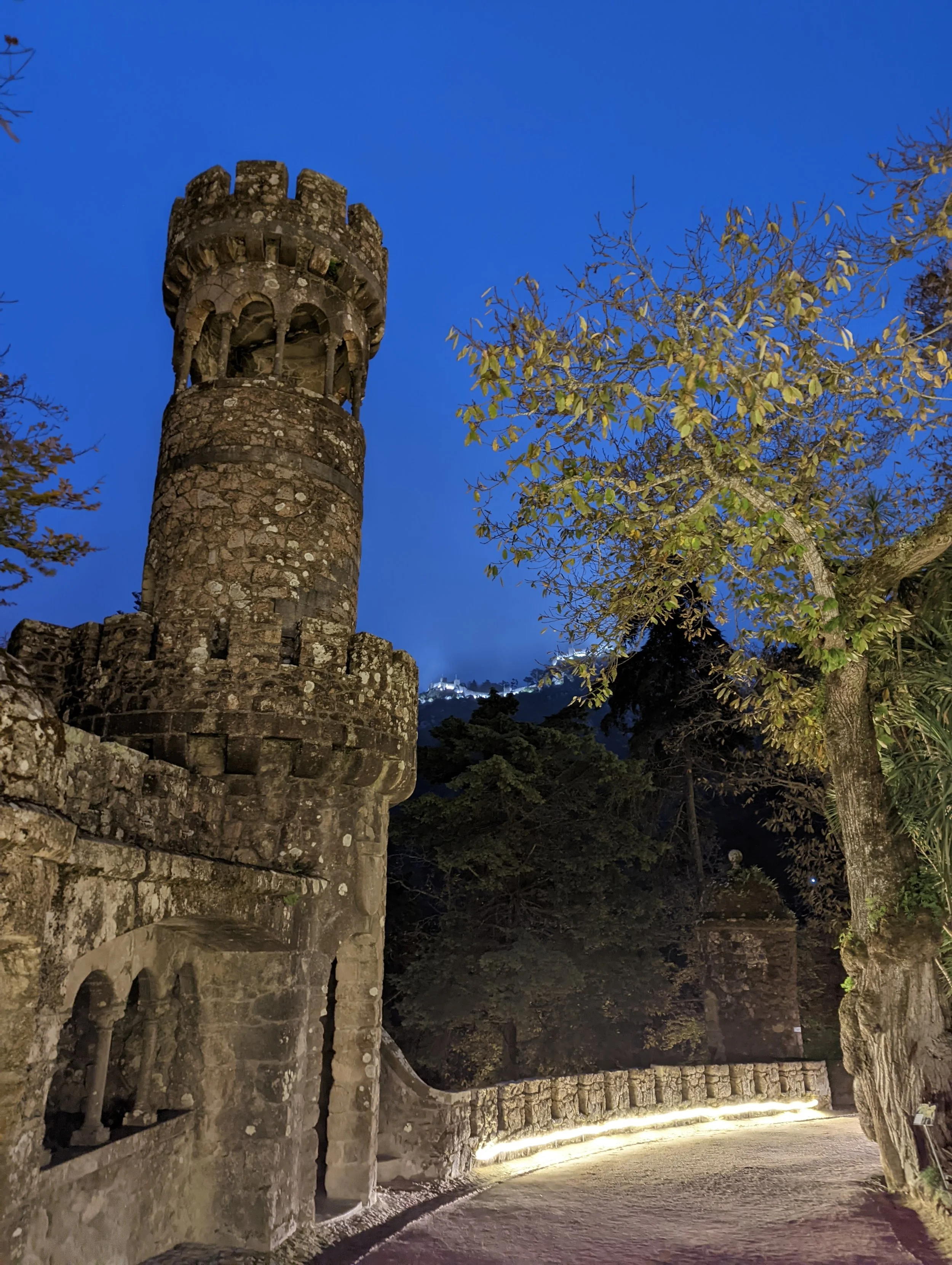 Night view of an old stone tower with battlements and arched openings, adjacent to a stone wall, with trees and illuminated pathway, under a dark blue sky.