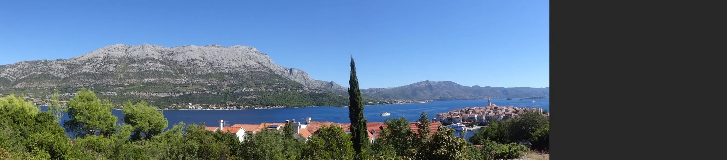Panoramic view of a coastal town with red rooftops, a large body of blue water, mountains in the background, a clear blue sky, and greenery in the foreground.