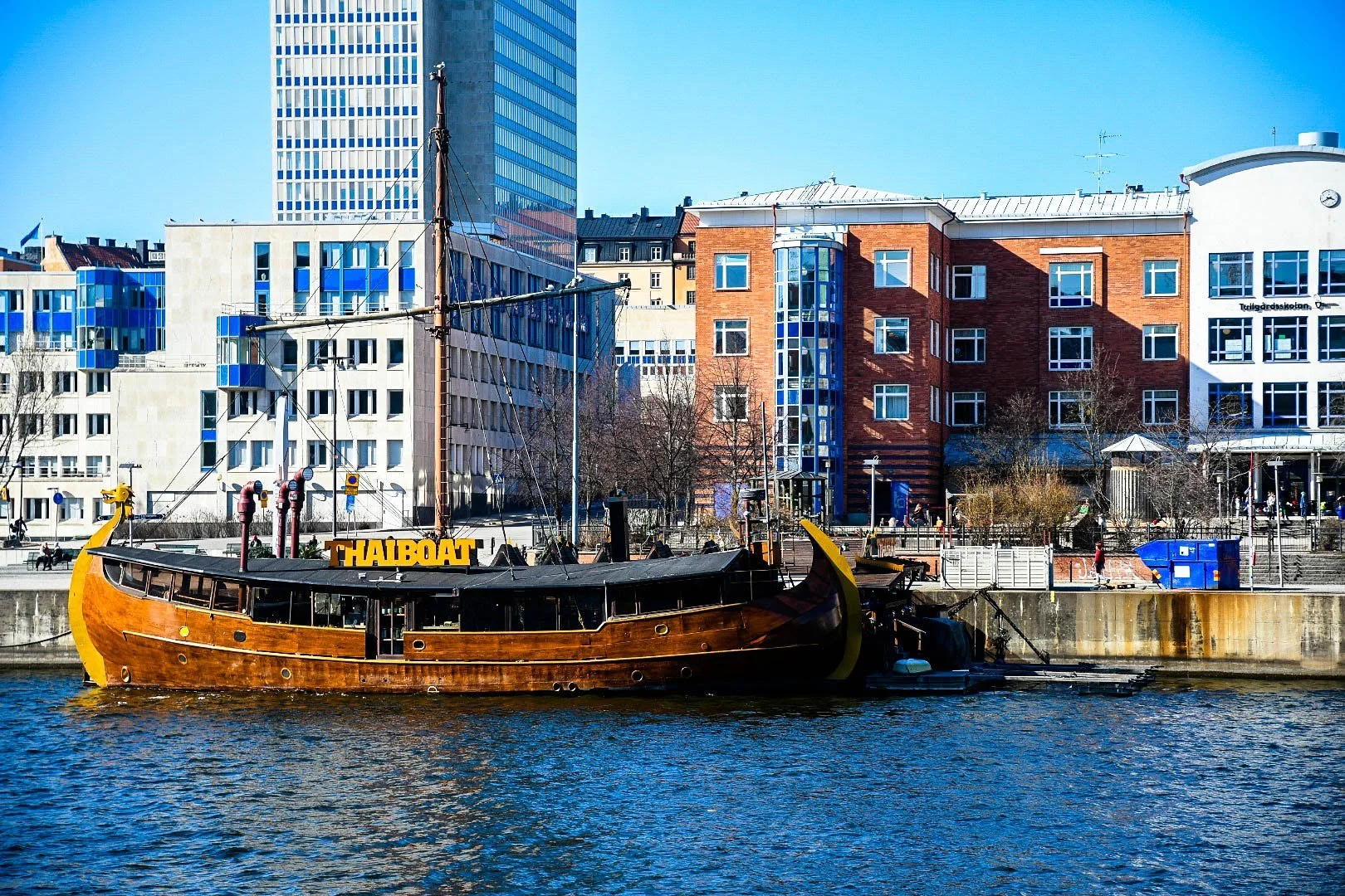 A wooden boat with the sign 'THAIBOAT' is docked along a waterfront with modern multi-story buildings in the background.