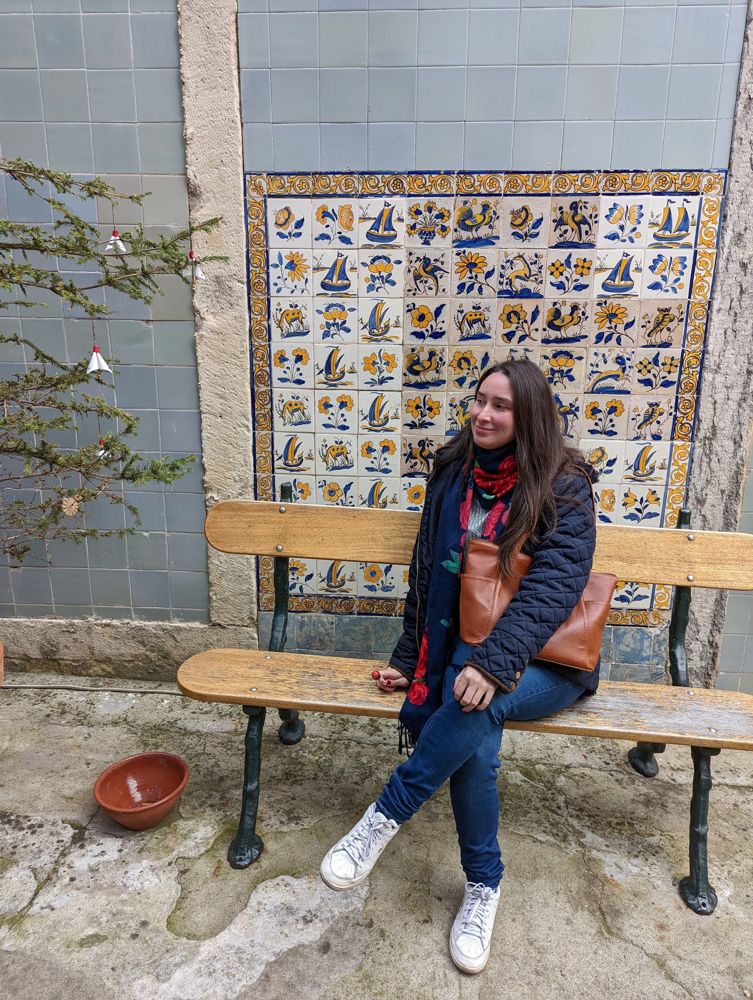 A woman with long dark hair, sitting on a wooden bench outdoors, wearing a navy quilted jacket, blue jeans, white sneakers, and a colorful scarf. She is holding a brown leather handbag. Behind her, there is a decorative wall of blue and yellow cerami
