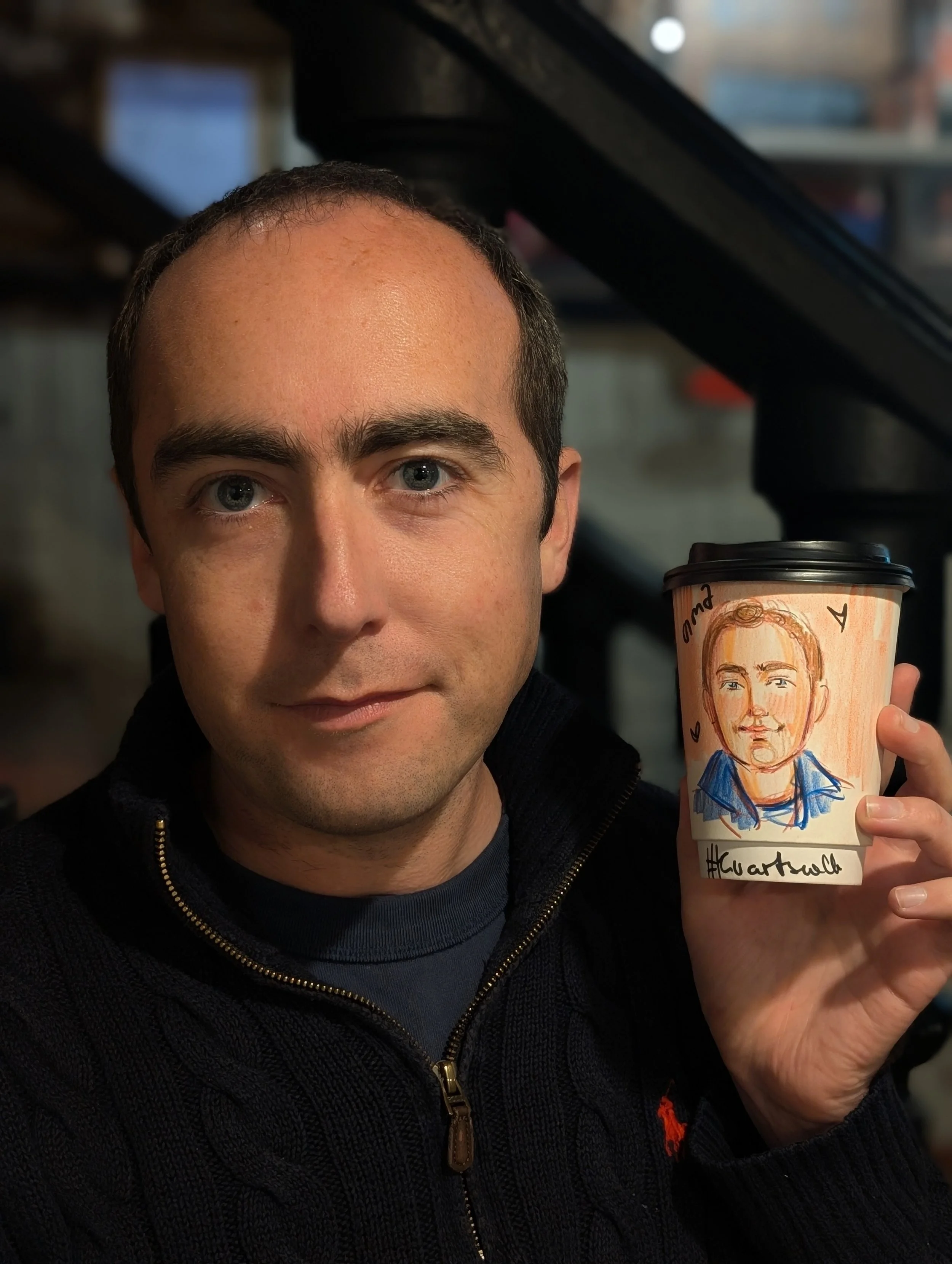 A man with short brown hair and blue eyes holding a coffee cup with a hand-drawn portrait of himself on it.