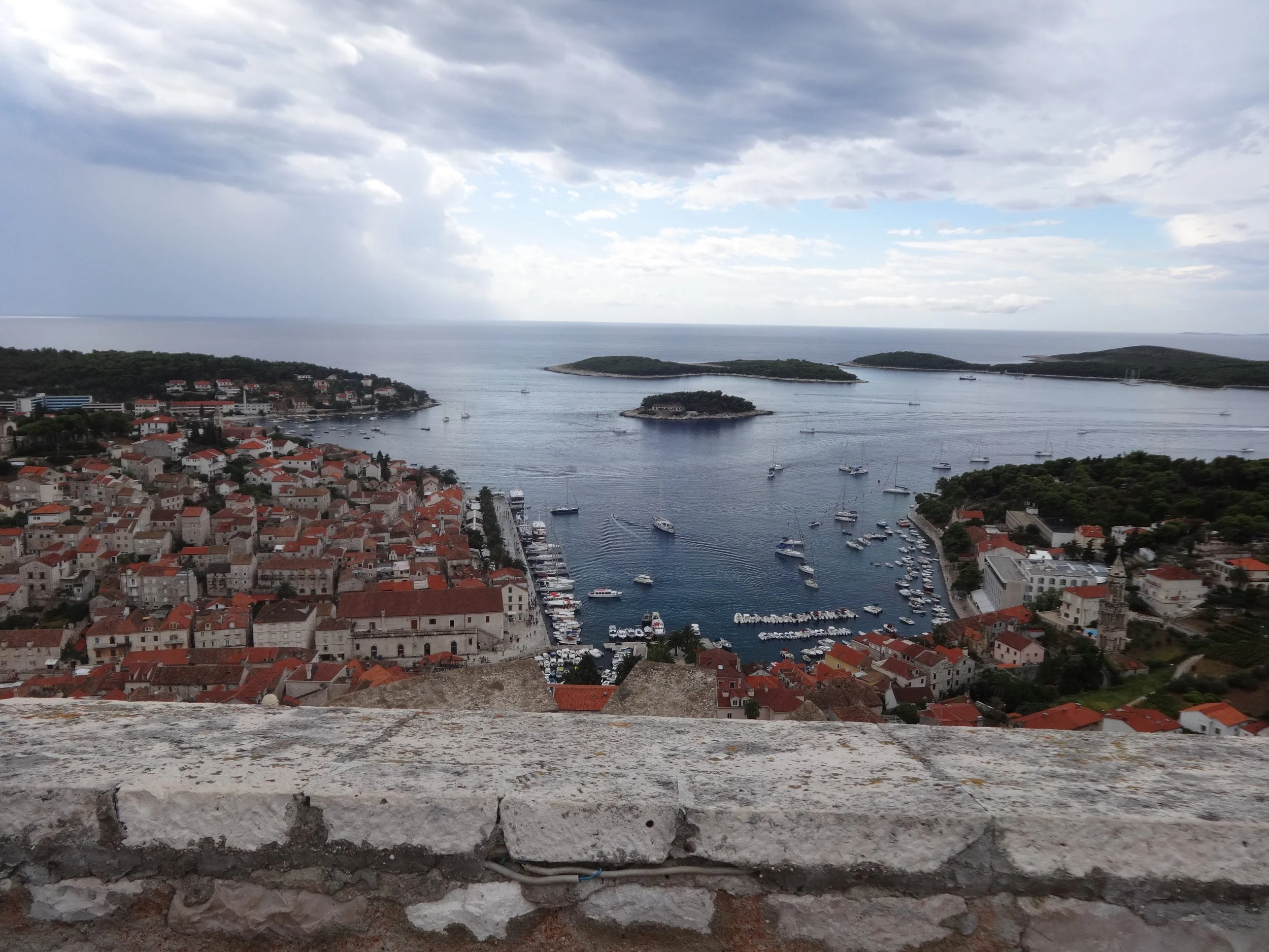 View of a coastal town with red-roofed buildings, a marina with boats, and green islands in the water under a cloudy sky.