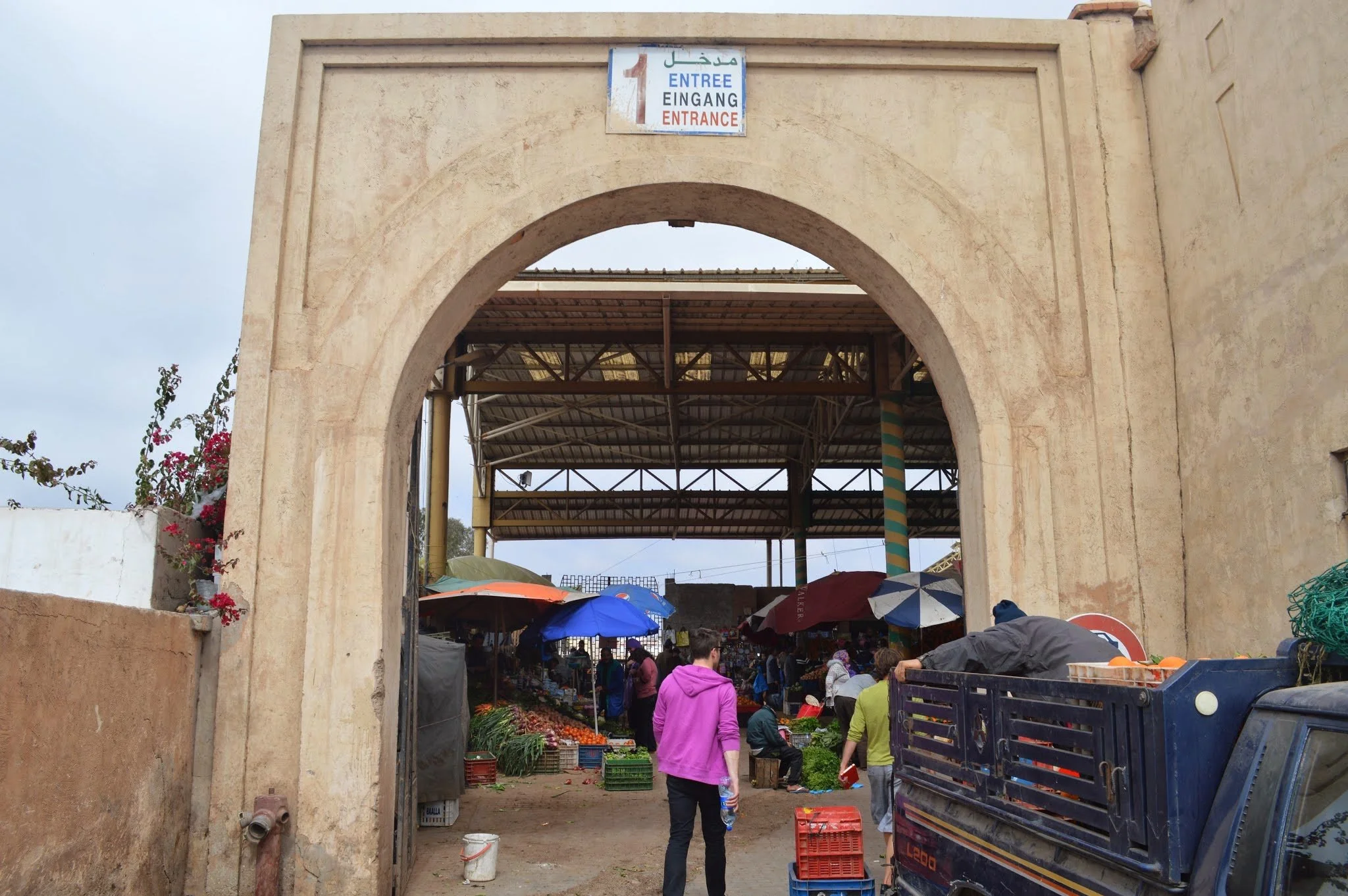 Entrance to an outdoor market with people shopping, under a large archway. Market stalls with umbrellas sell produce, and there is a sign in multiple languages indicating the entrance.