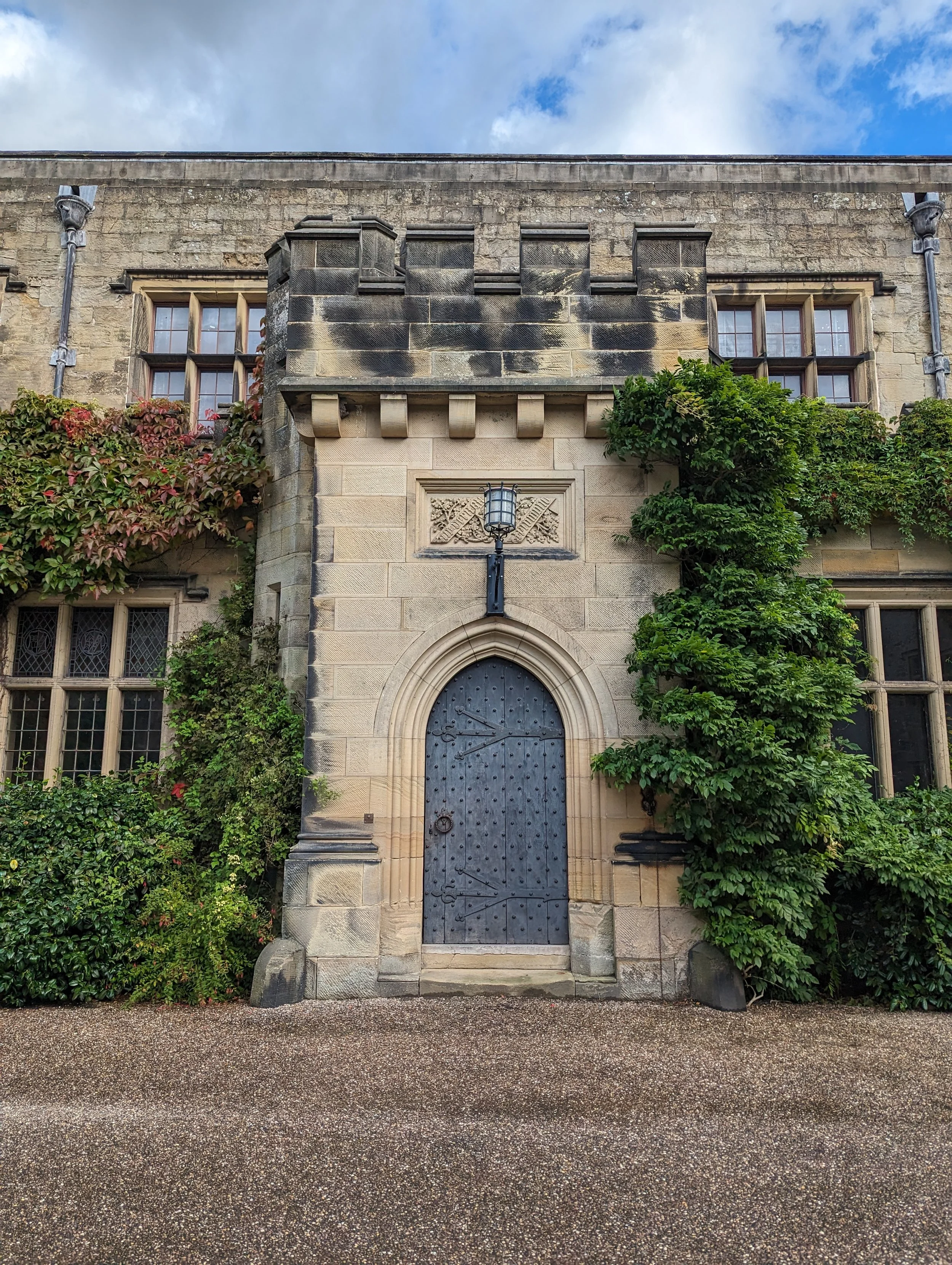 Front view of historic stone building with arched doorway, decorative stonework, ivy and shrubbery, and a lantern above the door.
