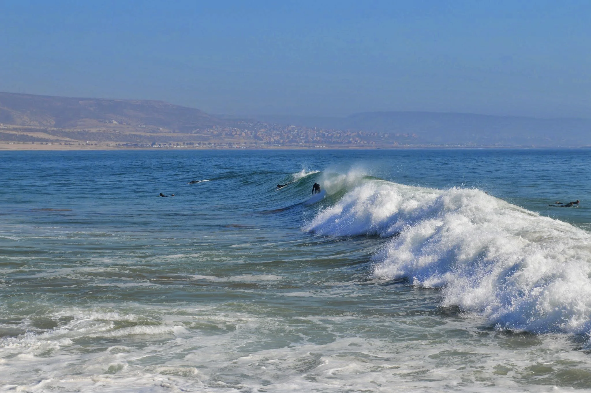 People surfing on ocean waves with a distant coastline and hills in the background on a clear day.
