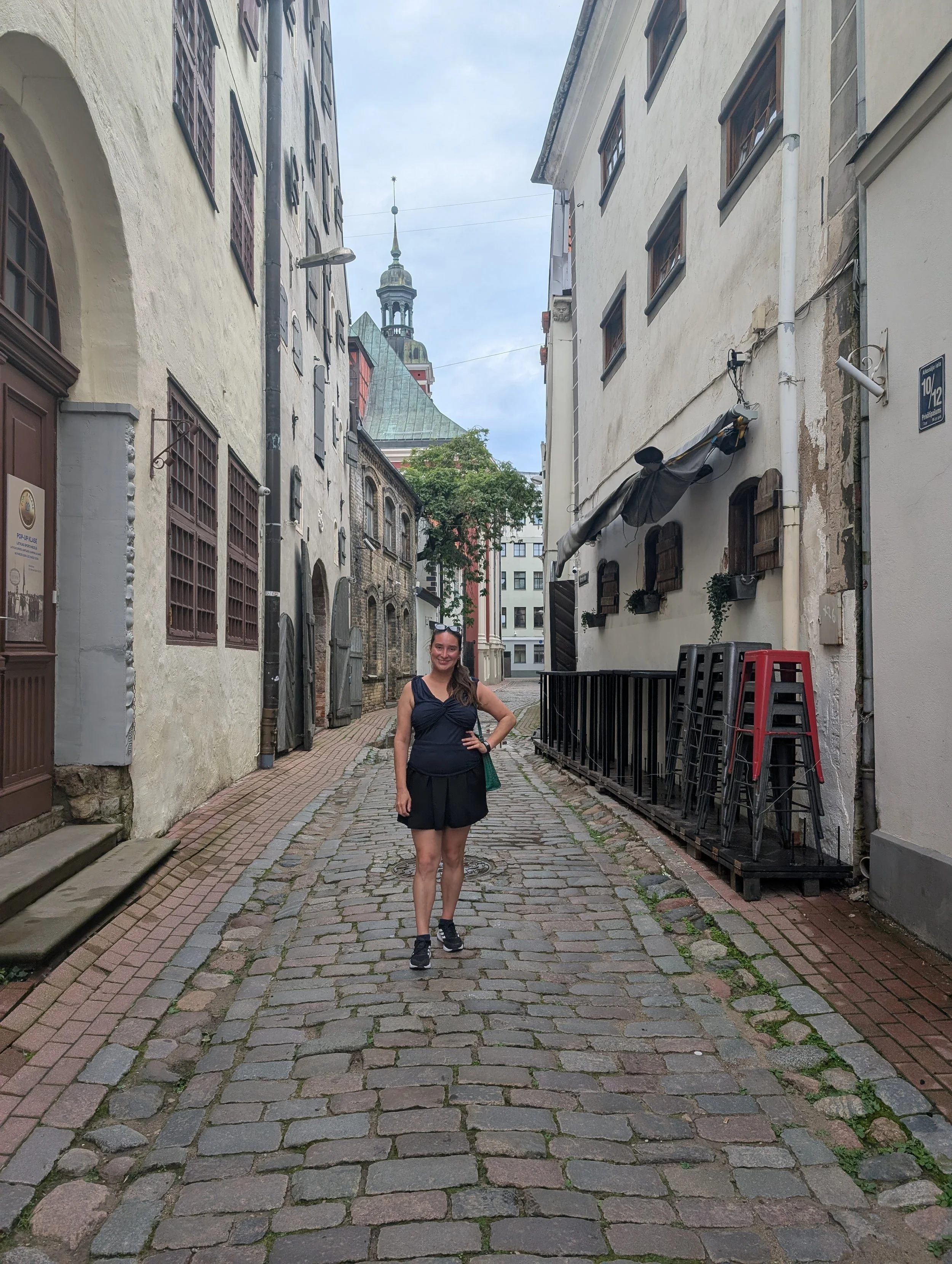 A woman standing on a cobblestone street in an alley with historic buildings on either side. She is smiling and wearing a black dress and sneakers, with a green bag on her shoulder. In the background, there is a church tower with a greenish spire.