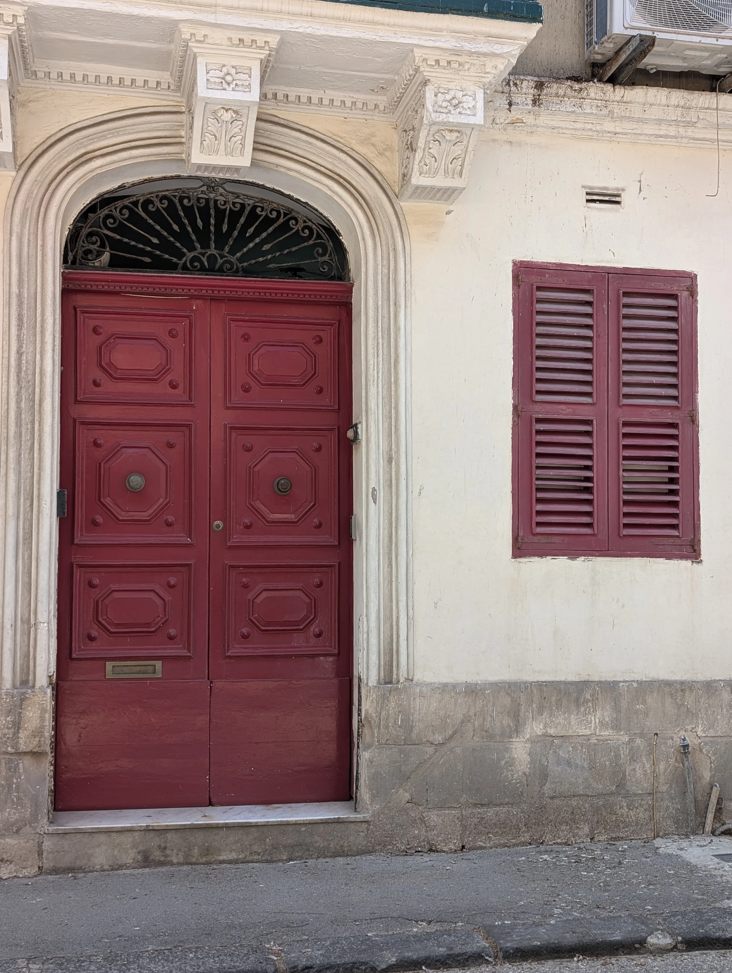 A red wooden door with decorative panels and a mail slot, set within an ornate stone archway, next to a window with red shutters, on a cream-colored building wall.