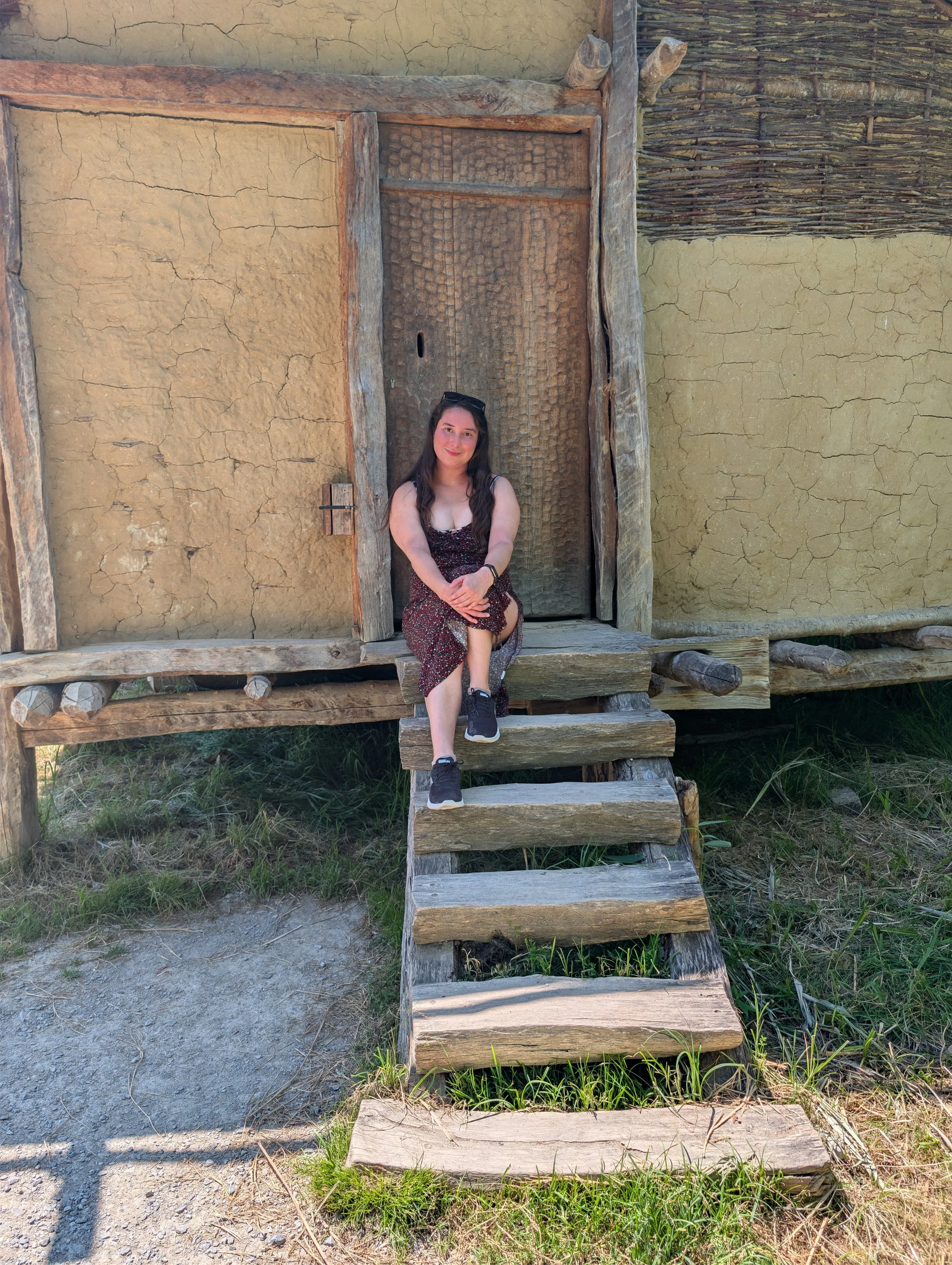A woman with long brown hair, wearing a floral dress and sneakers, sitting on weathered wooden steps outside a rustic building with mud walls and wooden accents. She is smiling, with sunglasses resting on her head, and arms crossed over a knee.