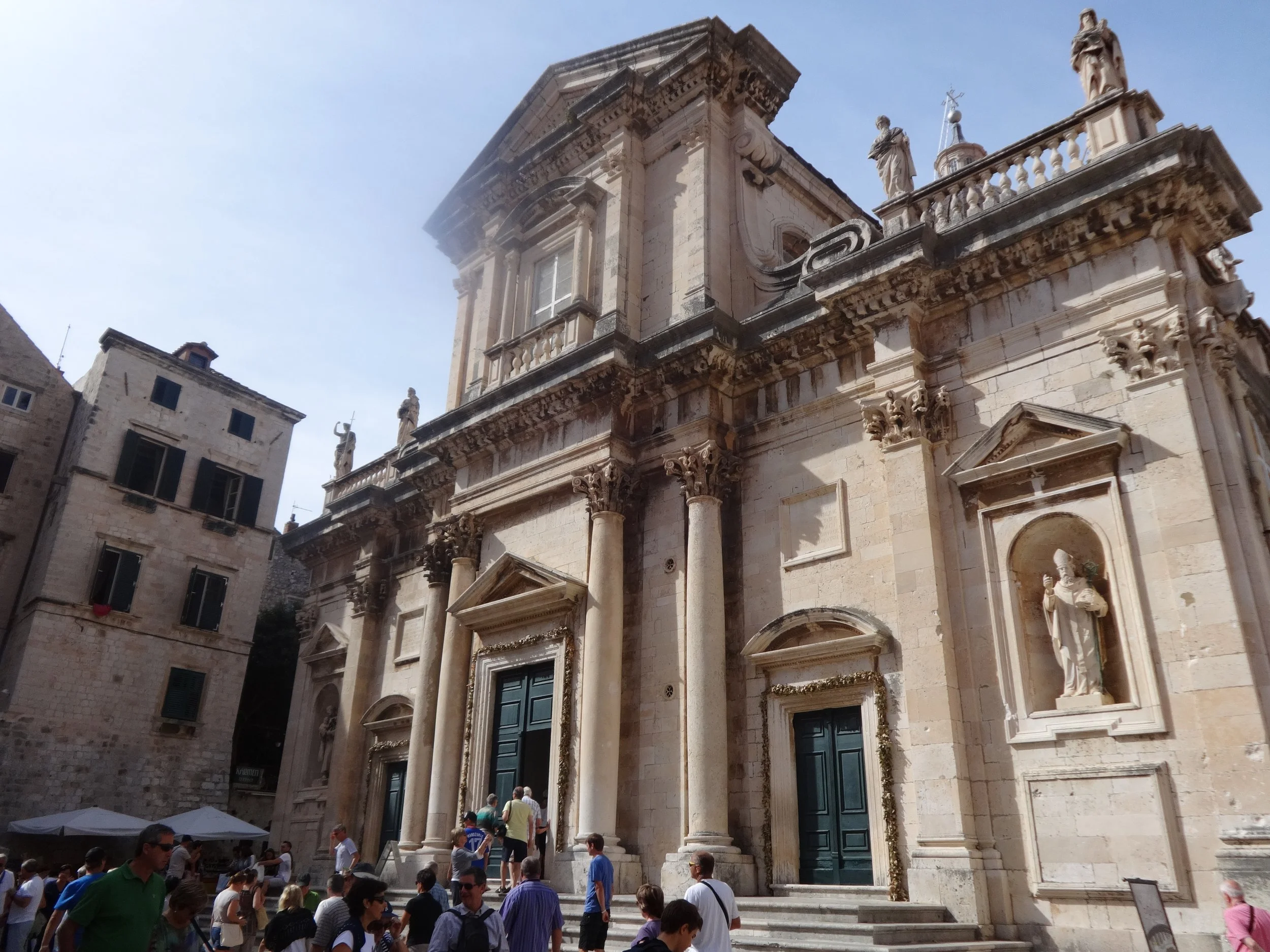 People gather outside a historic stone building with ornate columns and statues on the facade, likely a cathedral or church, under a clear blue sky.