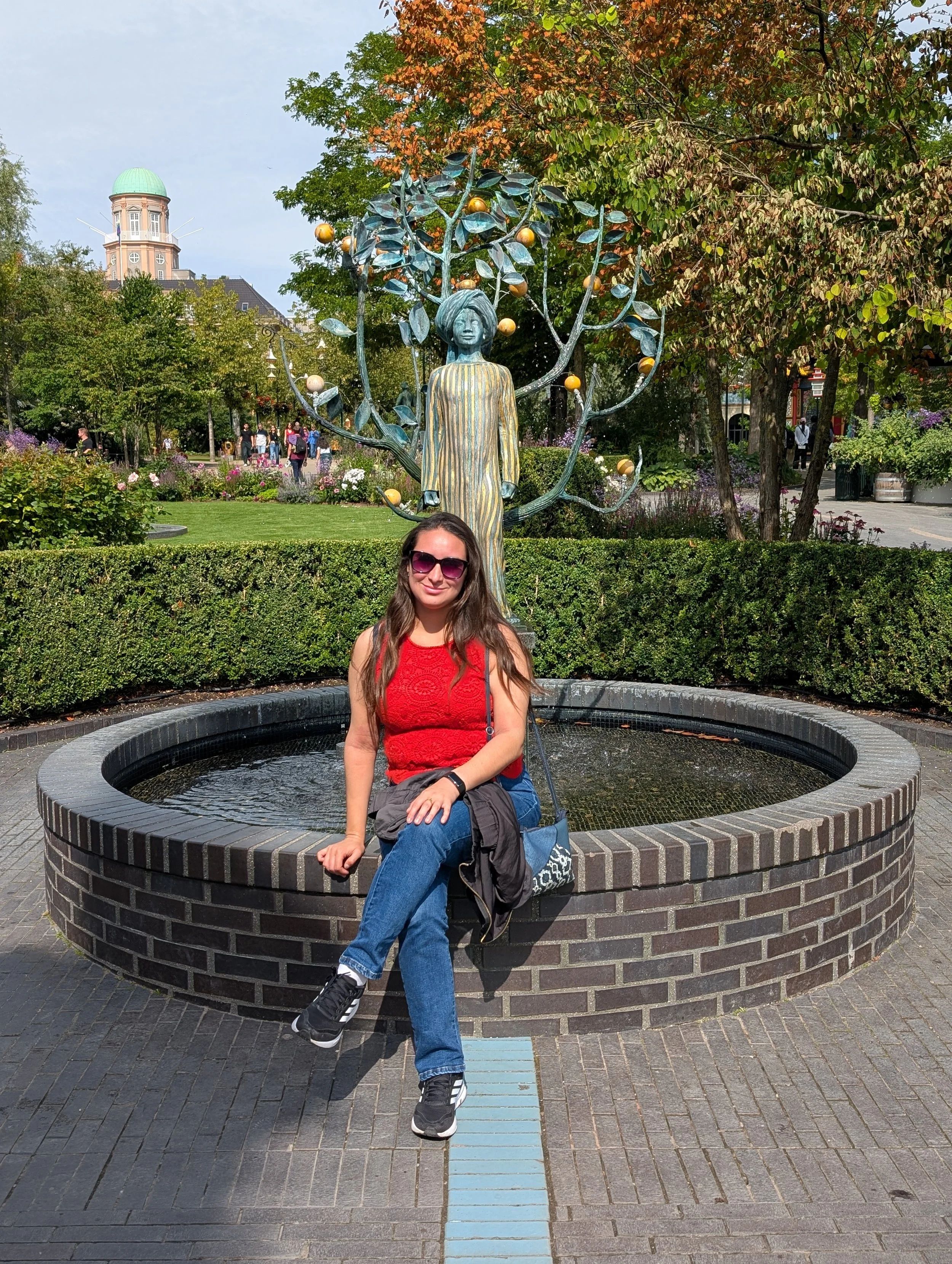 A woman sitting on a circular brick fountain in a park, with a young girl in a red dress and sunglasses in front. Behind them is a decorative metal sculpture of a tree with gold-colored balls, surrounded by greenery, trees, and a historic building wi