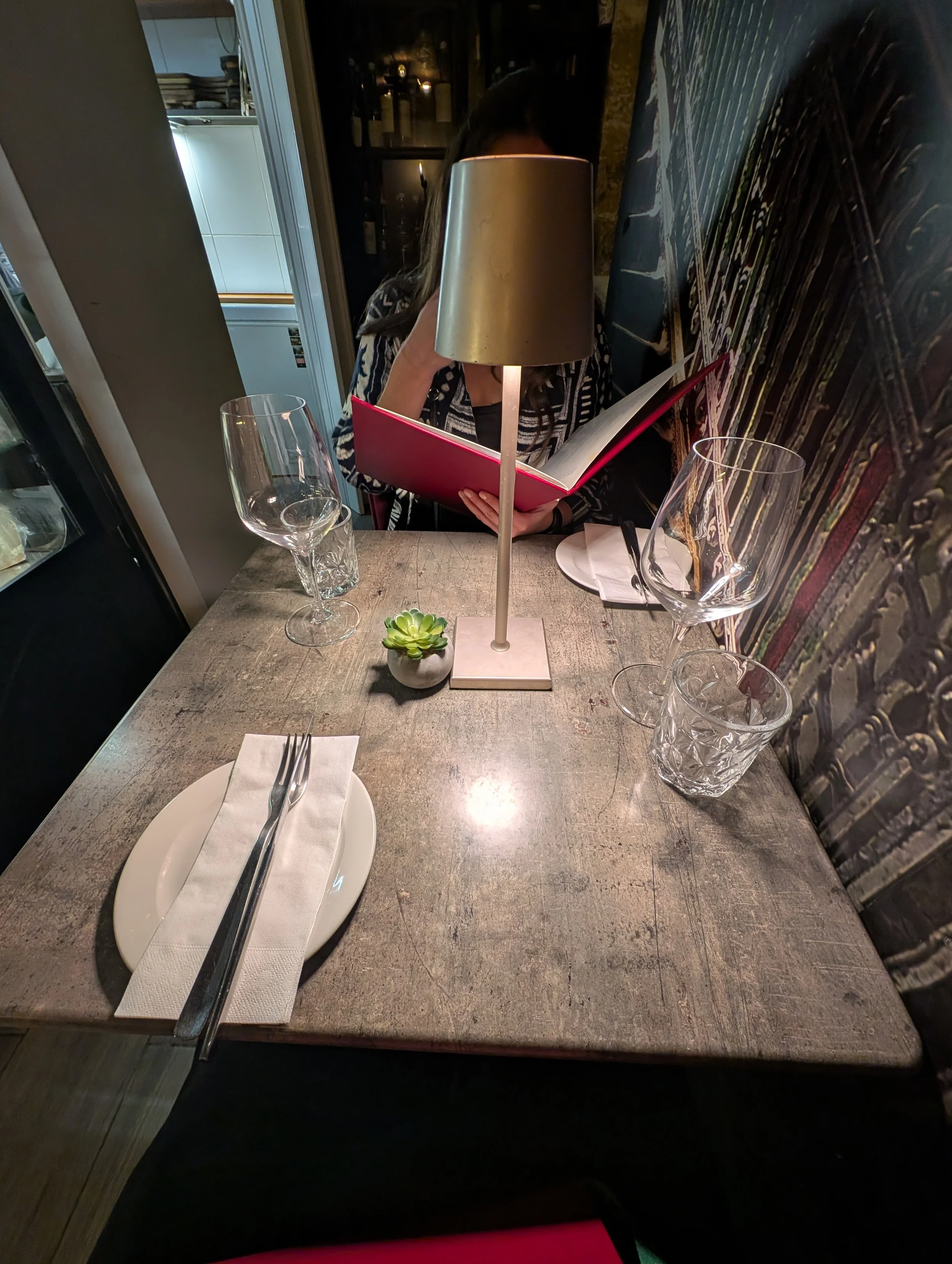 A person is sitting at a restaurant table reading a menu, with a lamp, empty wine glasses, a small plant, and cutlery on the table.