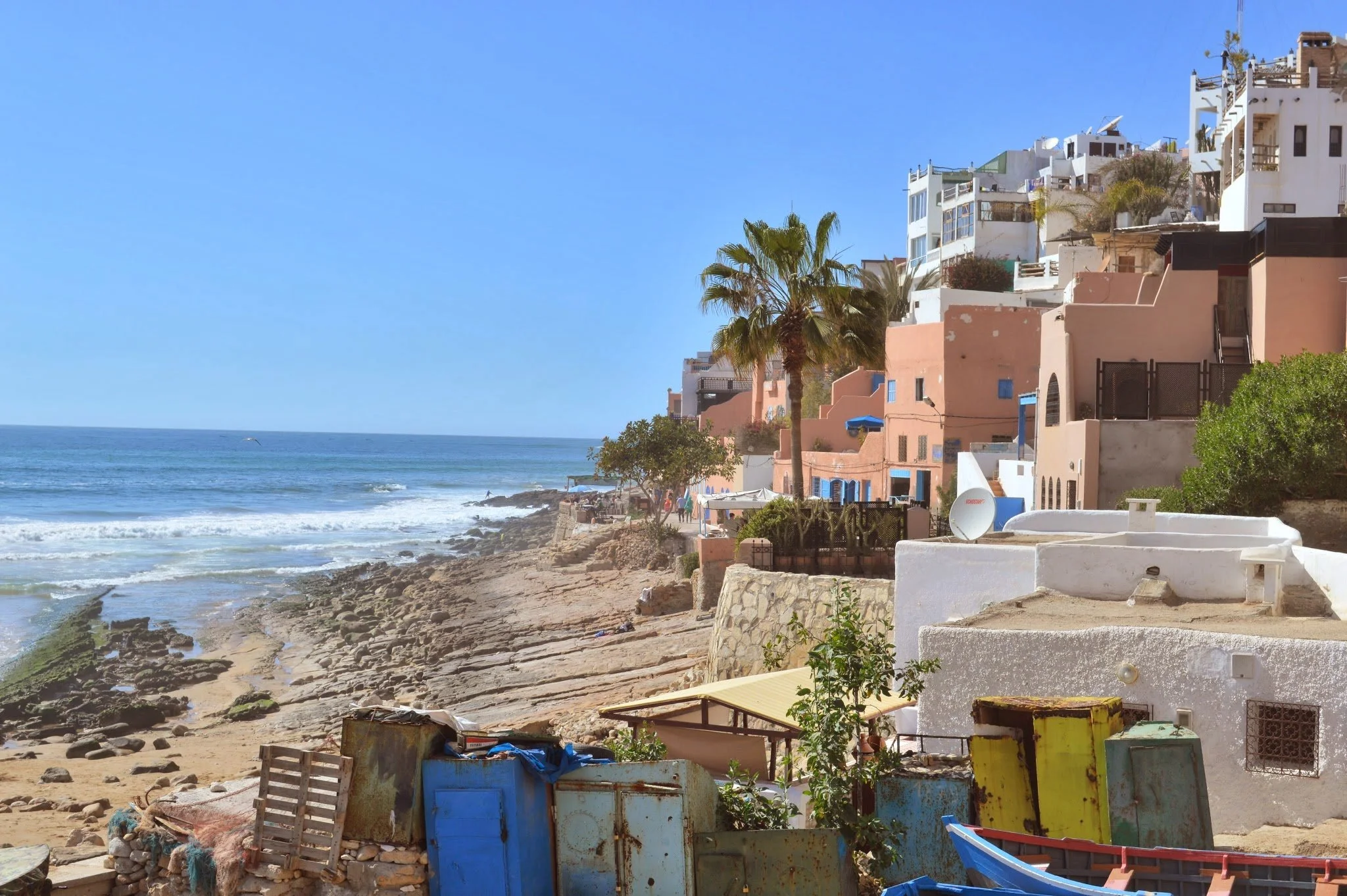 Houses with colourful facades on a hillside overlooking the ocean with waves and a clear blue sky, palm trees, and boats on the water in a coastal town.